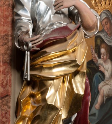 Zdjęcie nr 4: Rzeźba drążona z tyłu, ustawiona na niskiej podstawie. Figura ukazana frontalnie, z uniesioną głową, skierowaną w lewo, w kontrapoście, o esowato wygiętej sylwetce. Prawą dłoń z kluczami wyciąga w bok, lewą kładzie na piersi. Twarz pociągła o migdałowatych, blisko siebie umieszczonych oczach, lekko garbatym nosie i wąskich, rozchylonych ustach. Okolona jest krótką brodą oraz brązowymi, kręconymi włosami. Piotr ubrany jest w srebrzoną tunikę, przepasaną w talii, z kołnierzykiem oraz płaszcz przepasany w partii nóg, zawiązany na lewym biodrze, złocony o czerwonej podszewce. Polichromia ciała naturalistyczna. 
