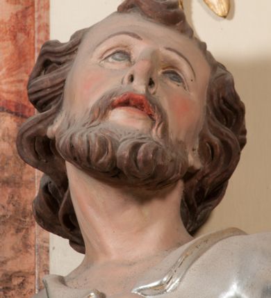 Zdjęcie nr 8: Rzeźba drążona z tyłu, ustawiona na niskiej podstawie. Figura ukazana frontalnie, z uniesioną głową, skierowaną w lewo, w kontrapoście, o esowato wygiętej sylwetce. Prawą dłoń z kluczami wyciąga w bok, lewą kładzie na piersi. Twarz pociągła o migdałowatych, blisko siebie umieszczonych oczach, lekko garbatym nosie i wąskich, rozchylonych ustach. Okolona jest krótką brodą oraz brązowymi, kręconymi włosami. Piotr ubrany jest w srebrzoną tunikę, przepasaną w talii, z kołnierzykiem oraz płaszcz przepasany w partii nóg, zawiązany na lewym biodrze, złocony o czerwonej podszewce. Polichromia ciała naturalistyczna. 
