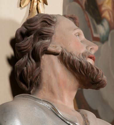 Zdjęcie nr 7: Rzeźba drążona z tyłu, ustawiona na niskiej podstawie. Figura ukazana frontalnie, z uniesioną głową, skierowaną w lewo, w kontrapoście, o esowato wygiętej sylwetce. Prawą dłoń z kluczami wyciąga w bok, lewą kładzie na piersi. Twarz pociągła o migdałowatych, blisko siebie umieszczonych oczach, lekko garbatym nosie i wąskich, rozchylonych ustach. Okolona jest krótką brodą oraz brązowymi, kręconymi włosami. Piotr ubrany jest w srebrzoną tunikę, przepasaną w talii, z kołnierzykiem oraz płaszcz przepasany w partii nóg, zawiązany na lewym biodrze, złocony o czerwonej podszewce. Polichromia ciała naturalistyczna. 
