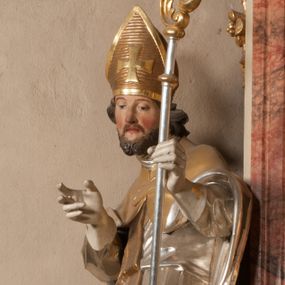 Zdjęcie nr 1: Święty zwrócony w trzech czwartych w lewo, o esowato wygiętej sylwetce. W lewej, ugiętej ręce trzyma pastorał, prawą wyciąga przed siebie. Twarz ma owalną, o okrągłych, półprzymkniętych oczach, uniesionych brwiach, lekko garbatym nosie i pełnych ustach o drążonych kącikach. Twarz ma okoloną krótką, kręconą brodą oraz silnie kręconymi, brązowymi włosami. Ubrany jest w czerwoną albę, srebrzoną rokietę o złoconej koronce przy brzegu oraz skrzyżowaną na piersi, złoconą stułę, przepasane w talii. Na ramiona ma narzuconą złoconą kapę ze srebrzoną podszewką, zapiętą klamrą na piersi, zawiniętą na prawej nodze. Na głowie ma złoconą infułę, na dłoniach białe rękawiczki. Po lewej stronie, u stóp św. Stanisława półpostać Piotrowina ukazanego tyłem do widza, unoszącego ręce w stronę świętego, z bandaną na głowie i piersiach. Polichromia w partiach ciała naturalistyczna. 



