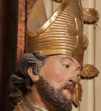 Zdjęcie nr 9: Święty zwrócony w trzech czwartych w lewo, o esowato wygiętej sylwetce. W lewej, ugiętej ręce trzyma pastorał, prawą wyciąga przed siebie. Twarz ma owalną, o okrągłych, półprzymkniętych oczach, uniesionych brwiach, lekko garbatym nosie i pełnych ustach o drążonych kącikach. Twarz ma okoloną krótką, kręconą brodą oraz silnie kręconymi, brązowymi włosami. Ubrany jest w czerwoną albę, srebrzoną rokietę o złoconej koronce przy brzegu oraz skrzyżowaną na piersi, złoconą stułę, przepasane w talii. Na ramiona ma narzuconą złoconą kapę ze srebrzoną podszewką, zapiętą klamrą na piersi, zawiniętą na prawej nodze. Na głowie ma złoconą infułę, na dłoniach białe rękawiczki. Po lewej stronie, u stóp św. Stanisława półpostać Piotrowina ukazanego tyłem do widza, unoszącego ręce w stronę świętego, z bandaną na głowie i piersiach. Polichromia w partiach ciała naturalistyczna. 



