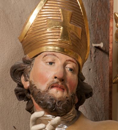 Zdjęcie nr 7: Święty zwrócony w trzech czwartych w lewo, o esowato wygiętej sylwetce. W lewej, ugiętej ręce trzyma pastorał, prawą wyciąga przed siebie. Twarz ma owalną, o okrągłych, półprzymkniętych oczach, uniesionych brwiach, lekko garbatym nosie i pełnych ustach o drążonych kącikach. Twarz ma okoloną krótką, kręconą brodą oraz silnie kręconymi, brązowymi włosami. Ubrany jest w czerwoną albę, srebrzoną rokietę o złoconej koronce przy brzegu oraz skrzyżowaną na piersi, złoconą stułę, przepasane w talii. Na ramiona ma narzuconą złoconą kapę ze srebrzoną podszewką, zapiętą klamrą na piersi, zawiniętą na prawej nodze. Na głowie ma złoconą infułę, na dłoniach białe rękawiczki. Po lewej stronie, u stóp św. Stanisława półpostać Piotrowina ukazanego tyłem do widza, unoszącego ręce w stronę świętego, z bandaną na głowie i piersiach. Polichromia w partiach ciała naturalistyczna. 



