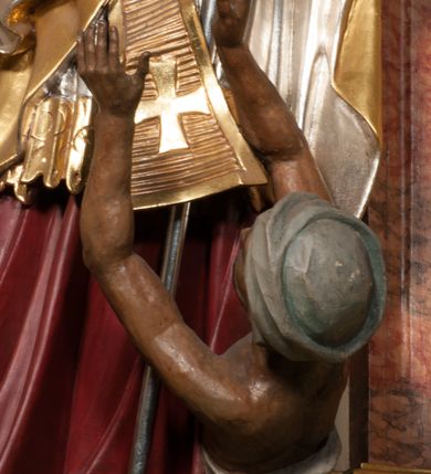 Zdjęcie nr 11: Święty zwrócony w trzech czwartych w lewo, o esowato wygiętej sylwetce. W lewej, ugiętej ręce trzyma pastorał, prawą wyciąga przed siebie. Twarz ma owalną, o okrągłych, półprzymkniętych oczach, uniesionych brwiach, lekko garbatym nosie i pełnych ustach o drążonych kącikach. Twarz ma okoloną krótką, kręconą brodą oraz silnie kręconymi, brązowymi włosami. Ubrany jest w czerwoną albę, srebrzoną rokietę o złoconej koronce przy brzegu oraz skrzyżowaną na piersi, złoconą stułę, przepasane w talii. Na ramiona ma narzuconą złoconą kapę ze srebrzoną podszewką, zapiętą klamrą na piersi, zawiniętą na prawej nodze. Na głowie ma złoconą infułę, na dłoniach białe rękawiczki. Po lewej stronie, u stóp św. Stanisława półpostać Piotrowina ukazanego tyłem do widza, unoszącego ręce w stronę świętego, z bandaną na głowie i piersiach. Polichromia w partiach ciała naturalistyczna. 



