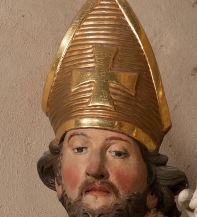 Zdjęcie nr 5: Święty zwrócony w trzech czwartych w lewo, o esowato wygiętej sylwetce. W lewej, ugiętej ręce trzyma pastorał, prawą wyciąga przed siebie. Twarz ma owalną, o okrągłych, półprzymkniętych oczach, uniesionych brwiach, lekko garbatym nosie i pełnych ustach o drążonych kącikach. Twarz ma okoloną krótką, kręconą brodą oraz silnie kręconymi, brązowymi włosami. Ubrany jest w czerwoną albę, srebrzoną rokietę o złoconej koronce przy brzegu oraz skrzyżowaną na piersi, złoconą stułę, przepasane w talii. Na ramiona ma narzuconą złoconą kapę ze srebrzoną podszewką, zapiętą klamrą na piersi, zawiniętą na prawej nodze. Na głowie ma złoconą infułę, na dłoniach białe rękawiczki. Po lewej stronie, u stóp św. Stanisława półpostać Piotrowina ukazanego tyłem do widza, unoszącego ręce w stronę świętego, z bandaną na głowie i piersiach. Polichromia w partiach ciała naturalistyczna. 



