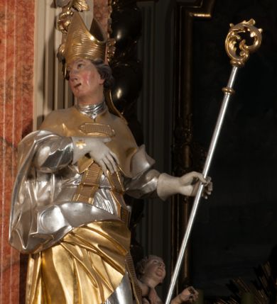 Zdjęcie nr 1: Święty ukazany frontalnie, z głową skierowaną w prawo, w kontrapoście, w lewej ręce trzyma pastorał, prawą kładzie na piersi. Twarz ma pociągłą, szczupłą, o podkreślonych kościach policzkowych i brodzie, o migdałowatych, głęboko rzeźbionych oczach, lekko zadartym nosie i rozchylonych ustach o drążonych kącikach, okoloną brązowymi, krótkimi, kręconymi włosami. Ubrany jest w czerwoną albę, srebrzoną rokietę ze złoconą koronką przy brzegu oraz złoconą stułę, przepasane w talii. Na ramiona ma narzuconą zapiętą pod szyją klamrą złoconą kapę o srebrzonej podszewce, zawiniętą na prawej nodze. Na głowie ma złoconą infułę, na dłoniach białe rękawiczki z krzyżykami. Po lewej stronie figury aniołek przepasany złoconą szarfą. Polichromia ciała naturalistyczna. 

