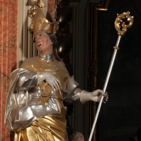 Zdjęcie nr 1: Święty ukazany frontalnie, z głową skierowaną w prawo, w kontrapoście, w lewej ręce trzyma pastorał, prawą kładzie na piersi. Twarz ma pociągłą, szczupłą, o podkreślonych kościach policzkowych i brodzie, o migdałowatych, głęboko rzeźbionych oczach, lekko zadartym nosie i rozchylonych ustach o drążonych kącikach, okoloną brązowymi, krótkimi, kręconymi włosami. Ubrany jest w czerwoną albę, srebrzoną rokietę ze złoconą koronką przy brzegu oraz złoconą stułę, przepasane w talii. Na ramiona ma narzuconą zapiętą pod szyją klamrą złoconą kapę o srebrzonej podszewce, zawiniętą na prawej nodze. Na głowie ma złoconą infułę, na dłoniach białe rękawiczki z krzyżykami. Po lewej stronie figury aniołek przepasany złoconą szarfą. Polichromia ciała naturalistyczna. 

