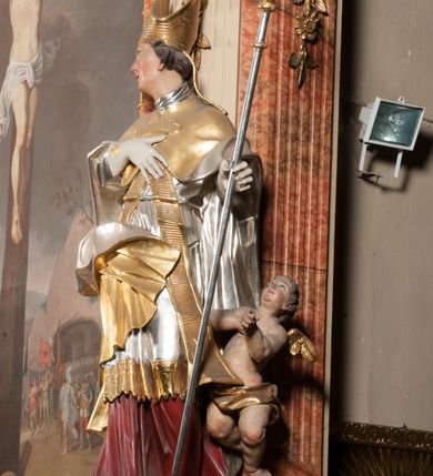 Zdjęcie nr 6: Święty ukazany frontalnie, z głową skierowaną w prawo, w kontrapoście, w lewej ręce trzyma pastorał, prawą kładzie na piersi. Twarz ma pociągłą, szczupłą, o podkreślonych kościach policzkowych i brodzie, o migdałowatych, głęboko rzeźbionych oczach, lekko zadartym nosie i rozchylonych ustach o drążonych kącikach, okoloną brązowymi, krótkimi, kręconymi włosami. Ubrany jest w czerwoną albę, srebrzoną rokietę ze złoconą koronką przy brzegu oraz złoconą stułę, przepasane w talii. Na ramiona ma narzuconą zapiętą pod szyją klamrą złoconą kapę o srebrzonej podszewce, zawiniętą na prawej nodze. Na głowie ma złoconą infułę, na dłoniach białe rękawiczki z krzyżykami. Po lewej stronie figury aniołek przepasany złoconą szarfą. Polichromia ciała naturalistyczna. 

