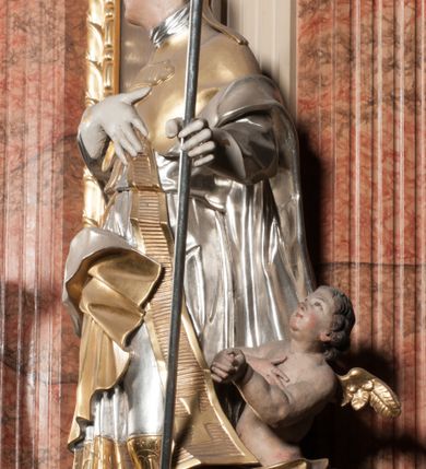 Zdjęcie nr 2: Święty ukazany frontalnie, z głową skierowaną w prawo, w kontrapoście, w lewej ręce trzyma pastorał, prawą kładzie na piersi. Twarz ma pociągłą, szczupłą, o podkreślonych kościach policzkowych i brodzie, o migdałowatych, głęboko rzeźbionych oczach, lekko zadartym nosie i rozchylonych ustach o drążonych kącikach, okoloną brązowymi, krótkimi, kręconymi włosami. Ubrany jest w czerwoną albę, srebrzoną rokietę ze złoconą koronką przy brzegu oraz złoconą stułę, przepasane w talii. Na ramiona ma narzuconą zapiętą pod szyją klamrą złoconą kapę o srebrzonej podszewce, zawiniętą na prawej nodze. Na głowie ma złoconą infułę, na dłoniach białe rękawiczki z krzyżykami. Po lewej stronie figury aniołek przepasany złoconą szarfą. Polichromia ciała naturalistyczna. 

