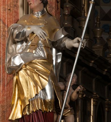 Zdjęcie nr 3: Święty ukazany frontalnie, z głową skierowaną w prawo, w kontrapoście, w lewej ręce trzyma pastorał, prawą kładzie na piersi. Twarz ma pociągłą, szczupłą, o podkreślonych kościach policzkowych i brodzie, o migdałowatych, głęboko rzeźbionych oczach, lekko zadartym nosie i rozchylonych ustach o drążonych kącikach, okoloną brązowymi, krótkimi, kręconymi włosami. Ubrany jest w czerwoną albę, srebrzoną rokietę ze złoconą koronką przy brzegu oraz złoconą stułę, przepasane w talii. Na ramiona ma narzuconą zapiętą pod szyją klamrą złoconą kapę o srebrzonej podszewce, zawiniętą na prawej nodze. Na głowie ma złoconą infułę, na dłoniach białe rękawiczki z krzyżykami. Po lewej stronie figury aniołek przepasany złoconą szarfą. Polichromia ciała naturalistyczna. 

