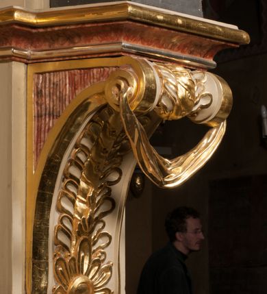 Zdjęcie nr 8: Święty ukazany frontalnie, z głową skierowaną w prawo, w kontrapoście, w lewej ręce trzyma pastorał, prawą kładzie na piersi. Twarz ma pociągłą, szczupłą, o podkreślonych kościach policzkowych i brodzie, o migdałowatych, głęboko rzeźbionych oczach, lekko zadartym nosie i rozchylonych ustach o drążonych kącikach, okoloną brązowymi, krótkimi, kręconymi włosami. Ubrany jest w czerwoną albę, srebrzoną rokietę ze złoconą koronką przy brzegu oraz złoconą stułę, przepasane w talii. Na ramiona ma narzuconą zapiętą pod szyją klamrą złoconą kapę o srebrzonej podszewce, zawiniętą na prawej nodze. Na głowie ma złoconą infułę, na dłoniach białe rękawiczki z krzyżykami. Po lewej stronie figury aniołek przepasany złoconą szarfą. Polichromia ciała naturalistyczna. 

