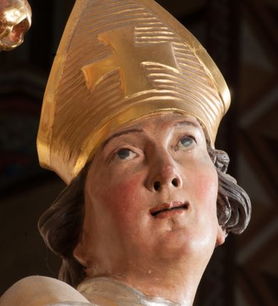 Zdjęcie nr 5: Święty ukazany frontalnie, z głową skierowaną w prawo, w kontrapoście, w lewej ręce trzyma pastorał, prawą kładzie na piersi. Twarz ma pociągłą, szczupłą, o podkreślonych kościach policzkowych i brodzie, o migdałowatych, głęboko rzeźbionych oczach, lekko zadartym nosie i rozchylonych ustach o drążonych kącikach, okoloną brązowymi, krótkimi, kręconymi włosami. Ubrany jest w czerwoną albę, srebrzoną rokietę ze złoconą koronką przy brzegu oraz złoconą stułę, przepasane w talii. Na ramiona ma narzuconą zapiętą pod szyją klamrą złoconą kapę o srebrzonej podszewce, zawiniętą na prawej nodze. Na głowie ma złoconą infułę, na dłoniach białe rękawiczki z krzyżykami. Po lewej stronie figury aniołek przepasany złoconą szarfą. Polichromia ciała naturalistyczna. 

