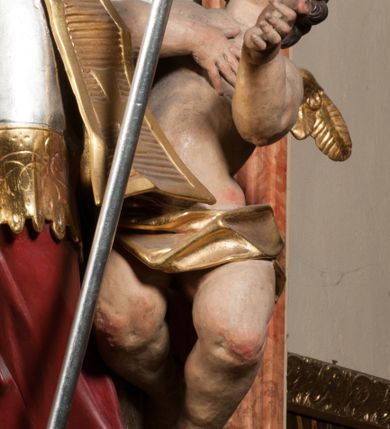 Zdjęcie nr 12: Święty ukazany frontalnie, z głową skierowaną w prawo, w kontrapoście, w lewej ręce trzyma pastorał, prawą kładzie na piersi. Twarz ma pociągłą, szczupłą, o podkreślonych kościach policzkowych i brodzie, o migdałowatych, głęboko rzeźbionych oczach, lekko zadartym nosie i rozchylonych ustach o drążonych kącikach, okoloną brązowymi, krótkimi, kręconymi włosami. Ubrany jest w czerwoną albę, srebrzoną rokietę ze złoconą koronką przy brzegu oraz złoconą stułę, przepasane w talii. Na ramiona ma narzuconą zapiętą pod szyją klamrą złoconą kapę o srebrzonej podszewce, zawiniętą na prawej nodze. Na głowie ma złoconą infułę, na dłoniach białe rękawiczki z krzyżykami. Po lewej stronie figury aniołek przepasany złoconą szarfą. Polichromia ciała naturalistyczna. 

