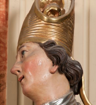 Zdjęcie nr 14: Święty ukazany frontalnie, z głową skierowaną w prawo, w kontrapoście, w lewej ręce trzyma pastorał, prawą kładzie na piersi. Twarz ma pociągłą, szczupłą, o podkreślonych kościach policzkowych i brodzie, o migdałowatych, głęboko rzeźbionych oczach, lekko zadartym nosie i rozchylonych ustach o drążonych kącikach, okoloną brązowymi, krótkimi, kręconymi włosami. Ubrany jest w czerwoną albę, srebrzoną rokietę ze złoconą koronką przy brzegu oraz złoconą stułę, przepasane w talii. Na ramiona ma narzuconą zapiętą pod szyją klamrą złoconą kapę o srebrzonej podszewce, zawiniętą na prawej nodze. Na głowie ma złoconą infułę, na dłoniach białe rękawiczki z krzyżykami. Po lewej stronie figury aniołek przepasany złoconą szarfą. Polichromia ciała naturalistyczna. 

