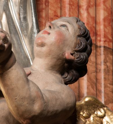 Zdjęcie nr 10: Święty ukazany frontalnie, z głową skierowaną w prawo, w kontrapoście, w lewej ręce trzyma pastorał, prawą kładzie na piersi. Twarz ma pociągłą, szczupłą, o podkreślonych kościach policzkowych i brodzie, o migdałowatych, głęboko rzeźbionych oczach, lekko zadartym nosie i rozchylonych ustach o drążonych kącikach, okoloną brązowymi, krótkimi, kręconymi włosami. Ubrany jest w czerwoną albę, srebrzoną rokietę ze złoconą koronką przy brzegu oraz złoconą stułę, przepasane w talii. Na ramiona ma narzuconą zapiętą pod szyją klamrą złoconą kapę o srebrzonej podszewce, zawiniętą na prawej nodze. Na głowie ma złoconą infułę, na dłoniach białe rękawiczki z krzyżykami. Po lewej stronie figury aniołek przepasany złoconą szarfą. Polichromia ciała naturalistyczna. 

