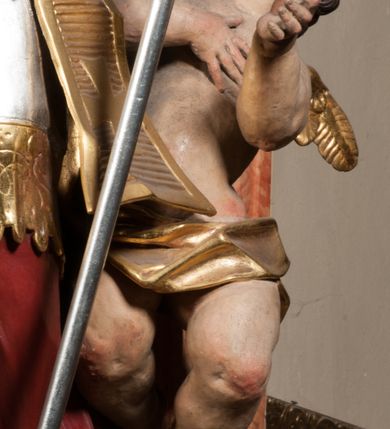 Zdjęcie nr 13: Święty ukazany frontalnie, z głową skierowaną w prawo, w kontrapoście, w lewej ręce trzyma pastorał, prawą kładzie na piersi. Twarz ma pociągłą, szczupłą, o podkreślonych kościach policzkowych i brodzie, o migdałowatych, głęboko rzeźbionych oczach, lekko zadartym nosie i rozchylonych ustach o drążonych kącikach, okoloną brązowymi, krótkimi, kręconymi włosami. Ubrany jest w czerwoną albę, srebrzoną rokietę ze złoconą koronką przy brzegu oraz złoconą stułę, przepasane w talii. Na ramiona ma narzuconą zapiętą pod szyją klamrą złoconą kapę o srebrzonej podszewce, zawiniętą na prawej nodze. Na głowie ma złoconą infułę, na dłoniach białe rękawiczki z krzyżykami. Po lewej stronie figury aniołek przepasany złoconą szarfą. Polichromia ciała naturalistyczna. 

