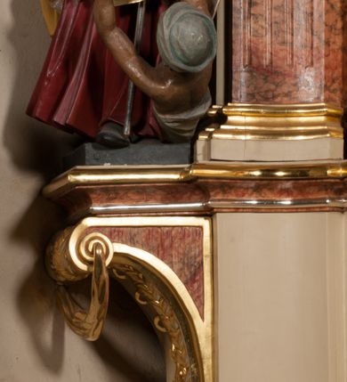 Zdjęcie nr 16: Święty ukazany frontalnie, z głową skierowaną w prawo, w kontrapoście, w lewej ręce trzyma pastorał, prawą kładzie na piersi. Twarz ma pociągłą, szczupłą, o podkreślonych kościach policzkowych i brodzie, o migdałowatych, głęboko rzeźbionych oczach, lekko zadartym nosie i rozchylonych ustach o drążonych kącikach, okoloną brązowymi, krótkimi, kręconymi włosami. Ubrany jest w czerwoną albę, srebrzoną rokietę ze złoconą koronką przy brzegu oraz złoconą stułę, przepasane w talii. Na ramiona ma narzuconą zapiętą pod szyją klamrą złoconą kapę o srebrzonej podszewce, zawiniętą na prawej nodze. Na głowie ma złoconą infułę, na dłoniach białe rękawiczki z krzyżykami. Po lewej stronie figury aniołek przepasany złoconą szarfą. Polichromia ciała naturalistyczna. 

