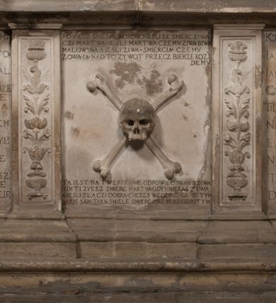 Zdjęcie nr 163: Nagrobek Grzegorza i Katarzyny Branickich znajduje się w kaplicy rodowej pod wezwaniem Najświętszej Marii Panny, na ścianie południowej, naprzeciwko wejścia. Składa się z trójosiowego cokołu i potrójnej filarowo-kolumnowej arkady z klęczącymi postaciami zmarłych po bokach adorujących znajdującą się pośrodku scenę ze zmartwychwstałym Chrystusem ukazującym się niewiernemu Tomaszowi. Całość wieńczy półkolista arkada z herbami Gryf i Kotwicz i tablicami inskrypcyjnymi. 
Dolna część artykułowa lizenami ozdobionymi motywami kandelabrowymi. W polu środkowym czaszka ze skrzyżowanymi piszczelami z inskrypcją w górnej i dolnej części: „POWIEDZ SVIETA [M]ADROSCI IESLI ZE SMIERC ZIWA / CZI MARTWA IESLI MARTWA CZEMV ZIWĄ BIWA / MALOWANA A IESLI ZIWA SMIERCIĄ CZEMV / ZOWIĄ IĄ NAD TO ZYWOT PRZECZ BIERZE KOZI/DEMV / TA IEST NA TWE PITANIE ODPOWIEC PRAWDZIWA / GDI TI ZYESZ SMIERC MARTVA GDY VMIERAS ZIWA / A IESLI ZLA CZI DOBRA CHCZES WIEDZIEC LI O TYM / IAKIS SĄM TAKĄ SMIELE SMIERC MIEĆ BĘDZIESZ PO TYM”. Na lewym polu inskrypcja „CO ZA DZIW ZEM POPIOLEM / WIEDZCIE ZESCIE WY / IAKO IA TENZE POPIOL, TO / IEDNO ZE ZYWY / BO GDIM SIĘ W PROCH ROSSIPAL / DOPIROM PRAWDZIWIE DOSZEDL, ZE CZLOWIEK TYMZE / PROCHEM CHOCIA ZYWIE / PATRZ ZE ZYWY POPIELIE A / TO POMNI SOBIE / ZES NA SWIECIE POPIOLEM / POPIOLEM I W GROBIE / A ZASZ NIEWIES CZYMEM BYL / A TERAZEM IAKI / OGLIANDAI, VWAZAIACZ ZE / SAM BEDZIESZ TAKI / BYLEM TERAS MIE NIE MASZ / IAK BIM TISZ NA SWIECIE / NIE BYL WY DZIS IESTESCIE / IUTRO NIEBEDZIECIE”. W polu cokołu „KTO TV LEZY POBOZNOSC / CNOTA OBYCZAIE / IAKICH NIERYCHLO VIZRĄ / BLISKO LEGLE KRAIE / IAKOSZ IE TAK MALY KĄT / MOGL ZACHOWAC W SOBIE / CZOSZ ALBO CNOTI LVCZKIE / GRZEBIĄ SCZIALEM W GROBIE / TILKOC TV CIALO LIEZY LIECZ / DVSZA W PRZESTRONE / NIEBIESKIE GMACHI WESŁA / KĘDI NIEZLICZONE / LATA POWIEDZIE. CZEMV IZE / MATKĄ BYLA / NIERZKAC POTOMSTWV ALIE / I SIROTOM SILA / TAKCI ZYWOT ZYWOTEM BYWA ZAKVPIONY / DOBRI FRIMARK DAC PLOCHI / ZA ON NIESKONCZONY”. Cokół zamyka wąski, silnie wysunięty gzyms z kimationem jońskim.
Trójosiowy podział nagrobka jest zachowany w partii środkowej, składającej się z trzech arkadowych wnęk, z których środkowa ujęta jest wolnostojącymi kolumnami jońskimi pokrytymi spiralnie ułożonymi pasami z motywem wici roślinnej, a sąsiednie od zewnątrz flankowane filarami pokrytymi motywami kandelabrowymi. W niezbyt głębokich niszach, których podniebienia zdobią kasetony z rozetami w kluczach uskrzydlone główki aniołków umieszczono wykonane w marmurze pińczowskim płaskorzeźby Chrystusa Zmartwychwstałego i adorujących go zmarłych, Grzegorza i Katarzynę Branickich. 
Zbawiciel jest ujęty całopostaciowo, frontalnie, w niewielkim wykroku, z głową zwróconą lekko do klęczącego po prawej stronie św. Tomasza. Jego ciało jest smukłe, wąskie w ramionach, z zaznaczonymi żebrami i mięśniami, osłonięte przewiązaną na biodrach szatą i płaszczem na lewym ramieniu. Chrystus ma pociągłą twarz, o wysokim czole, szerokich kościach policzkowych, wydatnym nosie i z głębokimi oczodołami. Przedstawiony został z długimi wąsami i brodą oraz opadającymi na ramiona, kręconymi włosami. W lewej ręce trzyma chorągiew i podtrzymuje szatę, a prawą rękę ma opuszczoną ku dołowi. 
Święty Tomasz ukazany jest z profilu, w szacie podwójnie przewiązanej w pasie sznurem i przerzuconym przez lewe ramie płaszczem. Twarz z brodą i długimi włosami, odwrócona w stronę Chrystusa. Do góry unosi również prawą rękę, zachowaną tylko do łokcia, lewą rękę trzyma ponad pasem. Figura mężczyzny wyrzeźbiona w kamieniu, polichromowana, przysłania wieniec z obłoków i uskrzydlonych głów puttów otaczający Chrystusa. 
Postacie stoją na podwójnym postumencie. W jego górnej części wyryto sentencję łacińską na prostokątnej tablicy umieszczonej w ozdobnym kartuszu, z zawiniętym spiralnie wycięciem krawędzi górnej i z dodaną w krawędzi dolnej główką uskrzydlonego putta. Napis na postumencie: „PER QVEM ET NOS / RESVRGERE DEVOTA / MENTE, CREDIMVS”. Na płycie poniżej płaskorzeźbione, leżące na prawym boku putto podpierające głowę na prawej ręce, natomiast w lewej trzymające klepsydrę. Nagie ciało, lekko przysłonięte draperią, spoczywa na poduszce z dwoma pomponami. Prawa noga jest wyprostowana, lewa zgięta w kolanie. Twarz pucołowata, z otwartymi oczami, otoczona gęstymi puklami loków. Przed dzieckiem leży czaszka. Nad nim znajduje się grawerowany kartusz z napisem: „HODIE MIHI / CRAS TIBI”. 
W arkadzie po lewej stronie przedstawiony został w trzech czwartych, zwrócony ramionami do widza Grzegorz Branicki. Postać klęczy na poduszce, szerokie dłonie złożone ma razem na piersiach, a głowę lekko uniesioną. Twarz Branickiego o pociągłych rysach, szerokich kościach policzkowych i dużych oczach, z wysokim czołem przysłoniętym zaczesanymi do przodu włosami. Ukazany z wąsami i długą, pofalowaną brodą. Ubrany jest w pełną, dekorowaną zbroję, spod której wystaje jaka i nogawice. Na szyi zawieszony łańcuch, a u stóp ostrogi. Przy nogach Branickiego leżą rękawice i hełm z trzema piórami. Nad głową wznoszą się panoplia, złożone z okrągłej tarczy i skrzyżowanych miecza, szabli, buławy, łuku, kołczanu ze strzałami i włóczni.
Po lewej stronie arkadę wypełnia ujęta z profilu, postać klęczącej na dwóch kolanach Katarzyny Branickiej. Jest ubrana w długą szubę, opadającą sztywnymi, rozchodzącymi się fałdami, u dołu załamującymi się, które zasłaniają sylwetkę. Na głowie ma czepiec przysłaniający włosy i podwikę. Twarz o łagodnych rysach, z długim nosem, małymi ustami i lekko zadartym podbródkiem. Ręce złożone razem na piersiach. Na palcach wskazującym i serdecznym lewej dłoni pierścienie, a ponad nadgarstkiem czterokrotnie nawinięty różaniec. Ponad figurą płaskorzeźbione tondo z Matką Boską z Dzieciątkiem otoczonymi wieńcem obłoków. Maria siedzi, ubrana w długą suknię, z welonem na głowie, z płaszczem zarzuconym na ramiona, którym lekko okrywa nagie Dzieciątko.
Zwieńczenie nagrobka o brzegach inkrustowanych marmurowymi kołami i elipsami, ujęte ornamentalnymi uszami, jest zakończone pasem z zawijającymi się do środkami kartuszami i równolegle ułożonymi liśćmi akantu. Tympanon wypełnia dekoracja złożona z czterech kartuszy: dwóch owalnych na tle paludamentu, zwieńczonych uskrzydloną główką anioła z godłami herbów Gryf i Kotwicz i dwóch podłużnych z inskrypcjami. Na tablicy po lewej stronie inskrypcja: „MAGNIFICO D(OMI)NO GREGORIO A RUSCA BRANICZKI OLIM VENA/TORI ET BURGRABIO CRAC(OVIENSI) CAPITANEO NIEPOŁOMIECEN(SI) VIRO / INTEGERRIMO DE REGIBVS SVIS ET RE PVB(LICA) BENE MERITO RELIGIO/NIS CATHOLICE AMANTISSIMO AMICIS FIDELISSIMO, OMNIBVS AFFABI/LI ET BENIGNO JOANNES A RUSCA BRANICZKI VENATOR CRACO(VIENSIS) CAPITANEVS NIEPOŁOMICEN(SI) MESTVS FILIUS PARENTI CHA/RISSIMO POSVIT OBYT 9 MAII A(NNO) D(OMINI) 1595 AETATIS SUE 60”. Napis wykuty po prawej stronie: “MAGNIFICAE D(OMI)NE CATHARINAE MAGNIFICI OLIM D(OMI)NI AUGUSTINI / KOTFICZ D(OMINI) SIGISMVNDI I POLONIAE REGIS STABVLI PREFECTI / VEXILIFERISQUE CURIE ET BURGRABY CRACO(VIENSIS) FILIE MATRONAE / LECTISSIME QVE CVM PVDORE ET MORVM MIRA ELEGANTIA / MARITI SUMMUM AMOREM VIRTUTE ET MODESTIA OMNIVM BENE/VOLENTIAM MERVISSET ACERBISSIMA MORTE AB OCVLIS EREPTA EST / IDEM FILIUS MATRI AMANTISSIME CVM LACRIMIS POSVIT. OBYT / 8 OCTOBRIS A(NNO) D)OMINI) 1588 AETATIS SUE 38". 
