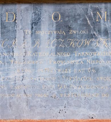 Zdjęcie nr 1: Płyta w formie leżącego prostokąta. W polu złocona inskrypcja 
„D(EO) O(PTIMO) M(AXIMO) / TU SPOCZYWAIĄ ZWŁOKI / JAKÓBA RACZKOWSKIEGO / KANONIKA KATEDRALNEGO TARNOWSKIEGO / DZIEKANA WIELICKIEGO PROBOSZCZA NIEPOŁOMSKIEGO / KTÓRY PRZEŻYWSZY LAT 64. / Z ŻALEM PARAFIANÓW I PRZYIACIOŁ SWOICH / POBOŻNE ZAKOŃCZYŁ ŻYCIE D(NIA) 6. PAŹDZIERNIKA R(OKU) 1816. / PRZECHODZĄCYCH PROSI O WESTCHNIENIE DO BOGA”.


