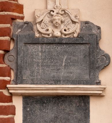 Zdjęcie nr 1: Tablica z wapienia dębnickiego w formie leżącego prostokąta ujętego grubą ramą, flankowaną falistymi uszakami z medalionami oraz z dekoracją w postaci dwóch cokolików z kulami przy górnej krawędzi. Tablica zwieńczona szczytem z wapienia pińczowskiego w formie kartusza utworzonego z ornamentu małżowinowo-chrząstkowego z uskrzydloną główką anielską w polu i guzem u dołu, zakończonego krzyżem o trójlistnych zakończeniach ramion z pięcioma rautami. Wzdłuż dolnej krawędzi fragment profilowanego gzymsu z wapienia pińczowskiego. Na górnym fragmencie ramy tablicy inskrypcja „I(ESU) M(ARIA) I(OSEPH)”, w polu zaś „CZYTAY NA ŚMIERĆ I DVSZE PAMIETAY / CO DEPCECIE PO TYM GROBIE / NASZYCH KOSCI MYSLCIE SOBIE / MIASTO IAKIEY KROTOFILI / BEDZIECIE Z NAMI PO CHWILI / TE DAIE KAMIEN PRZESTROGI / ZROWNA SIĘ PAN Y VBOGI. / ZMOWCIE PROSIEM POZDROWIENIE / DAY IEZV DVSZOM ZBAWIENIE”. Na dolnym fragmencie ramy inskrypcja „SEPVLTA OSSA DIE 24 8BPIS A(NNO) D(OMINI) 1697”.