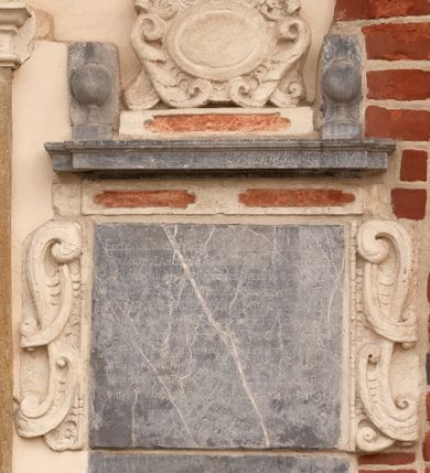 Zdjęcie nr 1: Tablica z wapienia dębnickiego w formie leżącego prostokąta. Flankowana uszakami z wapienia pińczowskiego utworzonymi z ornamentu małżowinowo-chrząstkowego. Wzdłuż górnej krawędzi płyty cokolik z wapienia pińczowskiego z dwiema płycinami polichromowanymi na czerwono. Ponad nim parapet z wapienia dębnickiego w formie gzymsu z dwoma wazonami, na nim mniejszy cokolik z wapienia pińczowskiego z płyciną polichromowaną na czerwono z kartuszem utworzonym z wolut i akantu, zwieńczony uskrzydloną główką anielską. W polu tablicy inskrypcja „D(EO) O(PTIMO) M(AXIMO) / ECCLE(SI)A HAEC EVDATA ET DOTATA PER SERENISS(IMO) CASIMIRVM AC / POLON(IAE) REGE(M) A(NNO) MCCCLIIII VETVSTATE AVTEM COLLAPSA REST/AVRATA PER ILL(USTRISSI)MOS RI PRINCIPES DVCES IN OSTROG ET / WISNIOWIEC COMITES IN WISNICZ IAROSŁAW ET TARNOW IOSEPH/VM CAROLVM ET THEOPHILAM LVBOMIRSCIOS MARE / SCHALCOS CVRIAE REGNI SANDOMIRIENSES NIEPOŁOM(ICENSES) ZA/TORIEN(SES) SOLECEN(SES) LUBACEN(SES) RICEN(SES) &amp; C(ETERA) CAPITANEOS ISTI/VS ECCL(ESI)AE BENEFACTORES NEC NON CVRAET SOLICITVDI/NE ADM(INISTARTORUM) R(EVERE)NDI(SSIMUS) THOMAE OLINSKI [C]VIVS PAROCHI AD FOR/MAM REDVCTATV QVI HAEC LECIS TANTIS BENEFACTO/RIBVS V TRAMQVE APPRECERE FELICITATEM A(NNO) D(OMINI) 1696”.