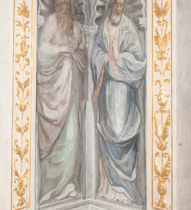 Zdjęcie nr 26: Iluzjonistyczną dekoracją malarską pokryto ściany kaplicy oraz strefę ponadcokołową: ściany tarczowe, pendentywy wokół stiukowych kartuszy herbowych oraz czaszę kopuły i wnętrze latarni. Ściany dolnej części podzielono na dwie strefy. Dolną tworzą zlokalizowane w narożach ścian iluzjonistycznie malowane pary nisz o konchach wypełnionych muszlą. W niszach ukazano osiem stojących frontalnie postaci starców, bez atrybutów i nimbów. Zapewne w ten sposób przedstawiono proroków lub apostołów. Każda z postaci ubrana jest w tunikę oraz płaszcz. Różnicuje je jedynie układ rąk, sposób kształtowania zarostu oraz kolorystyka szat. W drugiej strefie w przyłuczach arkad ukazano całopostaciowe przedstawienia uskrzydlonych aniołów trzymających narzędzia męki Pańskiej. Na ścianie wschodniej anioł prezentuje chustę Weroniki (z prawej) oraz latarnię (z lewej). Po bokach wejścia do kaplicy na ścianie północnej po prawej stronie anioł trzyma lancę i koronę cierniową, a po lewej krzyż. Z kolei na ścianie wschodniej po prawej ukazano anioła z drabiną, a po lewej z gąbką na trzcinie. Natomiast na ścianie południowej, po obu stronach zwieńczenia nagrobka, przedstawiono z prawej anioła z włócznią, a po lewej anioła z wiązką do biczowania. W tej strefie bezpośrednio przy łukach arkad wyobrażono leżące postacie geniuszy malowanych w technice en grisaille. Pod arkadą na ścianie wschodniej (ołtarzowej), ukazano dolną część owalnego jasno ugrowego baldachimu z lambrekinem i zielone, opadające paludamentum, rozsuwane kotarowo przez dwa uskrzydlone anioły. Dekoracja stanowi oprawę dla wiszącego tu obrazu Matki Boskiej z Dzieciątkiem w bogato rzeźbionej ramie. Z kolei na ścianie zachodniej wyobrażono monumentalną scenę Zdjęcia z krzyża. Jest to zwarta kompozycja ogniskująca się na wysokim drewnianym krzyżu, z którego ściągane jest martwe ciało Chrystusa. Z dwóch stron dłuższych ramion krzyż podtrzymują dwaj aniołowie. Ciało Jezusa opuszczane jest na białym pasie tkaniny przewiązanym pod jego ramionami. Chrystus ze zwisającą głową ma ręce ułożone wzdłuż ciała. Biodra przepasane ma białym perizonium. Jego skrzyżowane nogi są ugięte i jeszcze przybite gwoździem do krzyża. Gwóźdź wyciąga z nich obcęgami mężczyzna w niebieskiej, podwiniętej koszuli. Ciało opuszczają dwaj mężczyźni widoczni za poprzecznymi ramionami krzyża, trzymający końce taśmy. Człowiek z lewej strony w niebieskiej koszuli dodatkowo odbiera drugą białą taśmę od mężczyzny ubranego w szaroczarny strój. Osoba ta stoi na drabinie opartej o poprzeczne ramię krzyża. Drugi mężczyzna za krzyżem, w brązowej koszuli, lewą ręką trzyma koniec taśmy, a w uniesionej prawej młotek. Ciało Zbawiciela podtrzymuje lewą ręką kolejny mężczyzna stojący tyłem do widza na drugiej drabinie opartej o lewe ramię krzyża. Jest on ubrany w krótkie brązowe spodenki i bladozieloną koszulę. Pod krzyżem stoi zwarta siedmioosobowa grupa. Najprawdopodobniej z prawej strony ukazano św. Jana i Matkę Boską, na środku dwóch żołnierzy w ostro zakończonych hełmach, dwie kobiety, w tym zapewne Marię Magdalenę i starca widocznego z lewej strony kompozycji. Na ścianach tarczowych nad gzymsem wyobrażono po bokach okrągłych okien siedzących czterech ewangelistów i czterech ojców Kościoła. Na ścianie wschodniej znalazł się z prawej św. Jan z orłem, który w dziobie trzyma złoty kałamarz, a po lewej św. Mateusz rozmawiający z aniołem. Z kolei na ścianie południowej po lewej stronie przedstawiono św. Marka z lwem oraz św. Ambrożego trzymającego w prawej ręce naczynie z wylewającym się miodem. Na ścianie zachodniej z lewej znalazł się czytający św. Augustyn z sercem na dłoni, a z prawej św. Grzegorz Wielki z widoczną za nim tiarą i sową. Na ścianie północnej po lewej ukazano św. Hieronima z leżącym lwem, a po prawej stronie św. Łukasz z wołem. W pendentywach wykonano malarską dekorację w formie wijącej się wstęgi, na tle której widnieją stiukowe kartusze rollwerkowe, o owalnych kaboszonowych polach czwórdzielnych w krzyż. Po stronie wschodniej w pendentywach znajdują się dwa puste kartusze z niezachowanymi herbami. Natomiast herby zachowały się w pendentywach po stronie zachodniej. Na pendentywie północno-zachodnim: Jastrzębiec, Nowina, Gozdawa, Pobóg, a na południowo-zachodnim Gryf, Kotwicz, Kotwicz, Zabawa. W kopule wyobrażono Koronację Matki Boskiej adorowanej przez różnych świętych. Scenę koronacji Marii przez Trójcę Świętą przedstawiono w części wschodniej czaszy. Ukazana frontalnie Maria klęczy ze złożonymi rękami do modlitwy na obłokach. Ma owalną twarz okoloną jasnymi długimi włosami rozsypującymi się na jej ramiona. Na uniesionej ku górze głowie ma ubraną zsuwającą się białą chustę okrywającą włosy. Madonna ubrana jest w zasłaniający jej ramiona bladoniebieski płaszcz, opadający po bokach na obłoki. Nieznacznie wyżej z prawej strony na obłokach siedzi Bóg Ojciec. Wyobrażono go w trzech czwartych w prawo. Jest to starszy mężczyzna o siwych włosach i brodzie. W wyciągniętej prawej ręce nad głową Marii wraz z Chrystusem trzyma złotą koronę w typie zamkniętym. Z kolei w lewej podtrzymuje glob ze złotym krzyżykiem. Bóg Ojciec na głowie ma tiarę papieską. Ubrany jest w szaro-niebieską szatę oraz wzorzystą złotą kapę narzuconą na ramiona i spiętą pod szyją klamrą, z połami okrywającymi nogi. Naprzeciwko siedzi Chrystus widoczny w trzech czwartych w lewo z twarzą ukazaną w profilu. W wyciągniętej prawej ręce trzyma wraz z Bogiem Ojcem koronę, a lewą podtrzymuje złote berło. Chrystus ma jasne włosy opadające puklami na ramiona i podobną krótką brodę. Na głowie ma otwartą złotą koronę. Został przedstawiony z nagim torsem, na którym zaznaczono przebity bok. Jego postać otacza narzucony na ramiona bladoczerwony całun udrapowany w formie płaszcza. Nad koroną bliżej głowy Boga Ojca widoczna jest gołębica Ducha Świętego otoczona świetlistym otokiem. Ponad sceną koronacji wokół otworu do latarni wyobrażono wirujące złocisto-szare obłoki, na tle których widać uskrzydlone główki aniołków oraz postacie mniejszych i większych aniołków, które muzykują na różnych instrumentach, bądź też trzymają gałązki palmowe lub kwiaty albo kwiaty rozsypują. Uskrzydlone główki aniołków widoczne są także pośród obłoków, na których klęczy Madonna. Z prawej strony koronacji wyobrażono dwóch biskupów. Starszy, być może św. Wojciech, został przedstawiony w postawie półklęczącej w trzech czwartych w lewo. Prawą ręką zdaje się wskazywać na scenę koronacji, lewą zaś przyciska do piersi. Ma odkrytą głowę z widocznymi siwymi włosami i taką samą brodą. Ubrany jest w niebiesko-zielonkawą kapę ze złotym kapturem zakończonym chwostem. Na ziemi przy jego stopie widoczna jest złota infuła. Świętemu przyglądają się dwaj aniołowie widoczni z tyłu. Za nim klęczy młodszy biskup z leżącym przy nim na ziemi pastorałem i mieczem. Zapewne jest to św. Stanisław ze Szczepanowa. Święty ukazany został frontalnie z prawą ręką przyciśniętą do piersi, a z lewą nieznacznie odsuniętą w bok. Ma pociągłą twarz o krótkich jasnych włosach i podobnym zaroście. Ubrany jest w albę oraz szaro-bordową kapę o złotym kapturze. Przy jego kolanach stoi złota infuła, a za nim widoczny jest anioł z gałązką palmową. Za św. Stanisławem w rozwianych szatach klęczy św. Jadwiga Śląska, którą identyfikuje bryła kościoła widoczna przy jej stopach. Postać Jadwigi ukazano w profilu z założonymi rękami przed sobą. Na głowie ma niewielką złotą koronę, spod której widoczna jest biała chusta okrywająca włosy. Ubrana jest w bladoczerwoną suknię przepasaną w talii i narzucony na ramiona bladoszkarłatny płaszcz z gronostajowym kołnierzem i obszyciem. Za św. Jadwigą stoi inna święta z odsłoniętą lewą ręką wspartą o bok. Brak atrybutu nie pozwala na jej identyfikację. Ukazano ją w trzech czwartych w prawo z głową w profilu. Ma owalną twarz z wydatnym nosem. Na głowie ma niewielką złotą koronę, spod której widać jasną chustę okrywającą włosy. Ubrana jest w złocisto-szarą suknię z krótkimi rękawami i bladoczerwony rozwiany płaszcz. Pomiędzy obiema kobietami widać dwie postacie uskrzydlonych aniołów. Za świętymi siedzi z otwartą księgą wyobrażony frontalnie św. Jana Kanty. Jest to mężczyzna w średnim wieku z wysokim czołem, krótkimi brązowymi włosami i podobną brodą. Ubrany jest w bladofioletową togę akademicką z gronostajową peleryną. Trzymana przez niego księga ma czerwone okładki. Obok św. Jana Kantego namalowano czteroosobową grupę starszych mężczyzn. Pierwszy z nich, ujętych w trzech czwartych w prawo, zdaje się przyklękając, odbierać prawą ręką palmę męczeństwa podawaną mu przez widocznego nad nim anioła. Lewą ma ułożoną wzdłuż ciała. Ma podłużną twarz widoczną z profilu, otoczoną białymi włosami i spiczastą białą brodą. Ubrany jest w rozwianą jasnobeżową szatę przewiązaną w pasie i taki sam płaszcz narzucony na prawe ramię. Lewy rękaw tuniki jest podciągnięty, odsłaniając przedramię ręki. Na lewej nodze widoczny jest sandał. Za nim przedstawiono klęczącego w ujęciu trzech czwartych w lewo zakonnika eremitę, być może św. Pawła Pustelnika w białym habicie paulińskim. Zakonnik na wysokości bioder ma rozsunięte ręce. Jego twarz o wydatnym nosie otacza krzaczasta, długa, biała broda oraz białe włosy. W głębi za nim częściowo widać dwóch innych brodatych mężczyzn w beżowych szatach. Jeden z nich trzyma w dłoni palmę. W niewielkiej odległości za grupą mężczyzn na pierwszym planie klęczy na obłoku ukazany frontalnie św. Jacek. W rękach uniesionych na wysokości piersi trzyma wieżycową, złotą monstrancję i figurę Matki Boskiej. Dominikanin został przedstawiony z wysokim łysym czołem i wianuszkiem siwiejących włosów. Jego pociągła twarz o wyrazistym nosie i wąskich, nieznacznie zaciśniętych ustach pozbawiona jest zarostu. Ubrany jest w dominikański habit z czarnoniebieskim płaszczem. Z lewej strony sceny koronacji namalowano grupę czterech świętych. Na pierwszym planie widać klęczącego św. Stanisława Kostkę, wspartego na długim kiju z przywiązanym do niego węzełkiem. Święty został przedstawiony w profilu. Ma młodzieńczą, owalną twarz z krótkimi brązowymi włosami i rumianymi policzkami. Ubrany jest w bladobeżowy płaszcz, spod którego widać jasnoseledynową szatę. Za Stanisławem Kostką wyłania się w półpostaci św. Jan Nepomucen z drewnianym krzyżem podtrzymywanym lewą ręką. Jest to dojrzały mężczyzna o pociągłej twarzy, z brązowymi włosami i brodą. Z prawej strony św. Stanisława Kostki ukazano stojące dwie zakonnice, św. Kingę i zapewne bł. Salomeę. Kingę ukazano frontalnie w kontrapoście z głową uniesioną ku niebu w jasnobeżowym habicie klaryski wpadającym w brąz. Księżna prawą ręką przyciska do piersi bryłę soli, a w opuszczonej lewej trzyma kwiat lilii. U jej stóp widać model kościoła klasztornego w Starym Sączu. Obok stoi najprawdopodobniej bł. Salomea. Ukazano ją w mocnym kontrapoście, z dłońmi splecionymi na piersi i spojrzeniem skierowanym ku ziemi. Ubrana jest w rozwiany szaroniebieski habit i jasny welon. Wnętrze latarni wypełniają uskrzydlone głowy aniołków na tle złocisto-szarych obłoków.    







