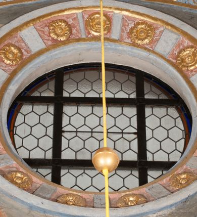 Zdjęcie nr 53: Iluzjonistyczną dekoracją malarską pokryto ściany kaplicy oraz strefę ponadcokołową: ściany tarczowe, pendentywy wokół stiukowych kartuszy herbowych oraz czaszę kopuły i wnętrze latarni. Ściany dolnej części podzielono na dwie strefy. Dolną tworzą zlokalizowane w narożach ścian iluzjonistycznie malowane pary nisz o konchach wypełnionych muszlą. W niszach ukazano osiem stojących frontalnie postaci starców, bez atrybutów i nimbów. Zapewne w ten sposób przedstawiono proroków lub apostołów. Każda z postaci ubrana jest w tunikę oraz płaszcz. Różnicuje je jedynie układ rąk, sposób kształtowania zarostu oraz kolorystyka szat. W drugiej strefie w przyłuczach arkad ukazano całopostaciowe przedstawienia uskrzydlonych aniołów trzymających narzędzia męki Pańskiej. Na ścianie wschodniej anioł prezentuje chustę Weroniki (z prawej) oraz latarnię (z lewej). Po bokach wejścia do kaplicy na ścianie północnej po prawej stronie anioł trzyma lancę i koronę cierniową, a po lewej krzyż. Z kolei na ścianie wschodniej po prawej ukazano anioła z drabiną, a po lewej z gąbką na trzcinie. Natomiast na ścianie południowej, po obu stronach zwieńczenia nagrobka, przedstawiono z prawej anioła z włócznią, a po lewej anioła z wiązką do biczowania. W tej strefie bezpośrednio przy łukach arkad wyobrażono leżące postacie geniuszy malowanych w technice en grisaille. Pod arkadą na ścianie wschodniej (ołtarzowej), ukazano dolną część owalnego jasno ugrowego baldachimu z lambrekinem i zielone, opadające paludamentum, rozsuwane kotarowo przez dwa uskrzydlone anioły. Dekoracja stanowi oprawę dla wiszącego tu obrazu Matki Boskiej z Dzieciątkiem w bogato rzeźbionej ramie. Z kolei na ścianie zachodniej wyobrażono monumentalną scenę Zdjęcia z krzyża. Jest to zwarta kompozycja ogniskująca się na wysokim drewnianym krzyżu, z którego ściągane jest martwe ciało Chrystusa. Z dwóch stron dłuższych ramion krzyż podtrzymują dwaj aniołowie. Ciało Jezusa opuszczane jest na białym pasie tkaniny przewiązanym pod jego ramionami. Chrystus ze zwisającą głową ma ręce ułożone wzdłuż ciała. Biodra przepasane ma białym perizonium. Jego skrzyżowane nogi są ugięte i jeszcze przybite gwoździem do krzyża. Gwóźdź wyciąga z nich obcęgami mężczyzna w niebieskiej, podwiniętej koszuli. Ciało opuszczają dwaj mężczyźni widoczni za poprzecznymi ramionami krzyża, trzymający końce taśmy. Człowiek z lewej strony w niebieskiej koszuli dodatkowo odbiera drugą białą taśmę od mężczyzny ubranego w szaroczarny strój. Osoba ta stoi na drabinie opartej o poprzeczne ramię krzyża. Drugi mężczyzna za krzyżem, w brązowej koszuli, lewą ręką trzyma koniec taśmy, a w uniesionej prawej młotek. Ciało Zbawiciela podtrzymuje lewą ręką kolejny mężczyzna stojący tyłem do widza na drugiej drabinie opartej o lewe ramię krzyża. Jest on ubrany w krótkie brązowe spodenki i bladozieloną koszulę. Pod krzyżem stoi zwarta siedmioosobowa grupa. Najprawdopodobniej z prawej strony ukazano św. Jana i Matkę Boską, na środku dwóch żołnierzy w ostro zakończonych hełmach, dwie kobiety, w tym zapewne Marię Magdalenę i starca widocznego z lewej strony kompozycji. Na ścianach tarczowych nad gzymsem wyobrażono po bokach okrągłych okien siedzących czterech ewangelistów i czterech ojców Kościoła. Na ścianie wschodniej znalazł się z prawej św. Jan z orłem, który w dziobie trzyma złoty kałamarz, a po lewej św. Mateusz rozmawiający z aniołem. Z kolei na ścianie południowej po lewej stronie przedstawiono św. Marka z lwem oraz św. Ambrożego trzymającego w prawej ręce naczynie z wylewającym się miodem. Na ścianie zachodniej z lewej znalazł się czytający św. Augustyn z sercem na dłoni, a z prawej św. Grzegorz Wielki z widoczną za nim tiarą i sową. Na ścianie północnej po lewej ukazano św. Hieronima z leżącym lwem, a po prawej stronie św. Łukasz z wołem. W pendentywach wykonano malarską dekorację w formie wijącej się wstęgi, na tle której widnieją stiukowe kartusze rollwerkowe, o owalnych kaboszonowych polach czwórdzielnych w krzyż. Po stronie wschodniej w pendentywach znajdują się dwa puste kartusze z niezachowanymi herbami. Natomiast herby zachowały się w pendentywach po stronie zachodniej. Na pendentywie północno-zachodnim: Jastrzębiec, Nowina, Gozdawa, Pobóg, a na południowo-zachodnim Gryf, Kotwicz, Kotwicz, Zabawa. W kopule wyobrażono Koronację Matki Boskiej adorowanej przez różnych świętych. Scenę koronacji Marii przez Trójcę Świętą przedstawiono w części wschodniej czaszy. Ukazana frontalnie Maria klęczy ze złożonymi rękami do modlitwy na obłokach. Ma owalną twarz okoloną jasnymi długimi włosami rozsypującymi się na jej ramiona. Na uniesionej ku górze głowie ma ubraną zsuwającą się białą chustę okrywającą włosy. Madonna ubrana jest w zasłaniający jej ramiona bladoniebieski płaszcz, opadający po bokach na obłoki. Nieznacznie wyżej z prawej strony na obłokach siedzi Bóg Ojciec. Wyobrażono go w trzech czwartych w prawo. Jest to starszy mężczyzna o siwych włosach i brodzie. W wyciągniętej prawej ręce nad głową Marii wraz z Chrystusem trzyma złotą koronę w typie zamkniętym. Z kolei w lewej podtrzymuje glob ze złotym krzyżykiem. Bóg Ojciec na głowie ma tiarę papieską. Ubrany jest w szaro-niebieską szatę oraz wzorzystą złotą kapę narzuconą na ramiona i spiętą pod szyją klamrą, z połami okrywającymi nogi. Naprzeciwko siedzi Chrystus widoczny w trzech czwartych w lewo z twarzą ukazaną w profilu. W wyciągniętej prawej ręce trzyma wraz z Bogiem Ojcem koronę, a lewą podtrzymuje złote berło. Chrystus ma jasne włosy opadające puklami na ramiona i podobną krótką brodę. Na głowie ma otwartą złotą koronę. Został przedstawiony z nagim torsem, na którym zaznaczono przebity bok. Jego postać otacza narzucony na ramiona bladoczerwony całun udrapowany w formie płaszcza. Nad koroną bliżej głowy Boga Ojca widoczna jest gołębica Ducha Świętego otoczona świetlistym otokiem. Ponad sceną koronacji wokół otworu do latarni wyobrażono wirujące złocisto-szare obłoki, na tle których widać uskrzydlone główki aniołków oraz postacie mniejszych i większych aniołków, które muzykują na różnych instrumentach, bądź też trzymają gałązki palmowe lub kwiaty albo kwiaty rozsypują. Uskrzydlone główki aniołków widoczne są także pośród obłoków, na których klęczy Madonna. Z prawej strony koronacji wyobrażono dwóch biskupów. Starszy, być może św. Wojciech, został przedstawiony w postawie półklęczącej w trzech czwartych w lewo. Prawą ręką zdaje się wskazywać na scenę koronacji, lewą zaś przyciska do piersi. Ma odkrytą głowę z widocznymi siwymi włosami i taką samą brodą. Ubrany jest w niebiesko-zielonkawą kapę ze złotym kapturem zakończonym chwostem. Na ziemi przy jego stopie widoczna jest złota infuła. Świętemu przyglądają się dwaj aniołowie widoczni z tyłu. Za nim klęczy młodszy biskup z leżącym przy nim na ziemi pastorałem i mieczem. Zapewne jest to św. Stanisław ze Szczepanowa. Święty ukazany został frontalnie z prawą ręką przyciśniętą do piersi, a z lewą nieznacznie odsuniętą w bok. Ma pociągłą twarz o krótkich jasnych włosach i podobnym zaroście. Ubrany jest w albę oraz szaro-bordową kapę o złotym kapturze. Przy jego kolanach stoi złota infuła, a za nim widoczny jest anioł z gałązką palmową. Za św. Stanisławem w rozwianych szatach klęczy św. Jadwiga Śląska, którą identyfikuje bryła kościoła widoczna przy jej stopach. Postać Jadwigi ukazano w profilu z założonymi rękami przed sobą. Na głowie ma niewielką złotą koronę, spod której widoczna jest biała chusta okrywająca włosy. Ubrana jest w bladoczerwoną suknię przepasaną w talii i narzucony na ramiona bladoszkarłatny płaszcz z gronostajowym kołnierzem i obszyciem. Za św. Jadwigą stoi inna święta z odsłoniętą lewą ręką wspartą o bok. Brak atrybutu nie pozwala na jej identyfikację. Ukazano ją w trzech czwartych w prawo z głową w profilu. Ma owalną twarz z wydatnym nosem. Na głowie ma niewielką złotą koronę, spod której widać jasną chustę okrywającą włosy. Ubrana jest w złocisto-szarą suknię z krótkimi rękawami i bladoczerwony rozwiany płaszcz. Pomiędzy obiema kobietami widać dwie postacie uskrzydlonych aniołów. Za świętymi siedzi z otwartą księgą wyobrażony frontalnie św. Jana Kanty. Jest to mężczyzna w średnim wieku z wysokim czołem, krótkimi brązowymi włosami i podobną brodą. Ubrany jest w bladofioletową togę akademicką z gronostajową peleryną. Trzymana przez niego księga ma czerwone okładki. Obok św. Jana Kantego namalowano czteroosobową grupę starszych mężczyzn. Pierwszy z nich, ujętych w trzech czwartych w prawo, zdaje się przyklękając, odbierać prawą ręką palmę męczeństwa podawaną mu przez widocznego nad nim anioła. Lewą ma ułożoną wzdłuż ciała. Ma podłużną twarz widoczną z profilu, otoczoną białymi włosami i spiczastą białą brodą. Ubrany jest w rozwianą jasnobeżową szatę przewiązaną w pasie i taki sam płaszcz narzucony na prawe ramię. Lewy rękaw tuniki jest podciągnięty, odsłaniając przedramię ręki. Na lewej nodze widoczny jest sandał. Za nim przedstawiono klęczącego w ujęciu trzech czwartych w lewo zakonnika eremitę, być może św. Pawła Pustelnika w białym habicie paulińskim. Zakonnik na wysokości bioder ma rozsunięte ręce. Jego twarz o wydatnym nosie otacza krzaczasta, długa, biała broda oraz białe włosy. W głębi za nim częściowo widać dwóch innych brodatych mężczyzn w beżowych szatach. Jeden z nich trzyma w dłoni palmę. W niewielkiej odległości za grupą mężczyzn na pierwszym planie klęczy na obłoku ukazany frontalnie św. Jacek. W rękach uniesionych na wysokości piersi trzyma wieżycową, złotą monstrancję i figurę Matki Boskiej. Dominikanin został przedstawiony z wysokim łysym czołem i wianuszkiem siwiejących włosów. Jego pociągła twarz o wyrazistym nosie i wąskich, nieznacznie zaciśniętych ustach pozbawiona jest zarostu. Ubrany jest w dominikański habit z czarnoniebieskim płaszczem. Z lewej strony sceny koronacji namalowano grupę czterech świętych. Na pierwszym planie widać klęczącego św. Stanisława Kostkę, wspartego na długim kiju z przywiązanym do niego węzełkiem. Święty został przedstawiony w profilu. Ma młodzieńczą, owalną twarz z krótkimi brązowymi włosami i rumianymi policzkami. Ubrany jest w bladobeżowy płaszcz, spod którego widać jasnoseledynową szatę. Za Stanisławem Kostką wyłania się w półpostaci św. Jan Nepomucen z drewnianym krzyżem podtrzymywanym lewą ręką. Jest to dojrzały mężczyzna o pociągłej twarzy, z brązowymi włosami i brodą. Z prawej strony św. Stanisława Kostki ukazano stojące dwie zakonnice, św. Kingę i zapewne bł. Salomeę. Kingę ukazano frontalnie w kontrapoście z głową uniesioną ku niebu w jasnobeżowym habicie klaryski wpadającym w brąz. Księżna prawą ręką przyciska do piersi bryłę soli, a w opuszczonej lewej trzyma kwiat lilii. U jej stóp widać model kościoła klasztornego w Starym Sączu. Obok stoi najprawdopodobniej bł. Salomea. Ukazano ją w mocnym kontrapoście, z dłońmi splecionymi na piersi i spojrzeniem skierowanym ku ziemi. Ubrana jest w rozwiany szaroniebieski habit i jasny welon. Wnętrze latarni wypełniają uskrzydlone głowy aniołków na tle złocisto-szarych obłoków.    







