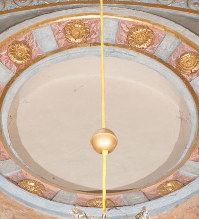 Zdjęcie nr 69: Iluzjonistyczną dekoracją malarską pokryto ściany kaplicy oraz strefę ponadcokołową: ściany tarczowe, pendentywy wokół stiukowych kartuszy herbowych oraz czaszę kopuły i wnętrze latarni. Ściany dolnej części podzielono na dwie strefy. Dolną tworzą zlokalizowane w narożach ścian iluzjonistycznie malowane pary nisz o konchach wypełnionych muszlą. W niszach ukazano osiem stojących frontalnie postaci starców, bez atrybutów i nimbów. Zapewne w ten sposób przedstawiono proroków lub apostołów. Każda z postaci ubrana jest w tunikę oraz płaszcz. Różnicuje je jedynie układ rąk, sposób kształtowania zarostu oraz kolorystyka szat. W drugiej strefie w przyłuczach arkad ukazano całopostaciowe przedstawienia uskrzydlonych aniołów trzymających narzędzia męki Pańskiej. Na ścianie wschodniej anioł prezentuje chustę Weroniki (z prawej) oraz latarnię (z lewej). Po bokach wejścia do kaplicy na ścianie północnej po prawej stronie anioł trzyma lancę i koronę cierniową, a po lewej krzyż. Z kolei na ścianie wschodniej po prawej ukazano anioła z drabiną, a po lewej z gąbką na trzcinie. Natomiast na ścianie południowej, po obu stronach zwieńczenia nagrobka, przedstawiono z prawej anioła z włócznią, a po lewej anioła z wiązką do biczowania. W tej strefie bezpośrednio przy łukach arkad wyobrażono leżące postacie geniuszy malowanych w technice en grisaille. Pod arkadą na ścianie wschodniej (ołtarzowej), ukazano dolną część owalnego jasno ugrowego baldachimu z lambrekinem i zielone, opadające paludamentum, rozsuwane kotarowo przez dwa uskrzydlone anioły. Dekoracja stanowi oprawę dla wiszącego tu obrazu Matki Boskiej z Dzieciątkiem w bogato rzeźbionej ramie. Z kolei na ścianie zachodniej wyobrażono monumentalną scenę Zdjęcia z krzyża. Jest to zwarta kompozycja ogniskująca się na wysokim drewnianym krzyżu, z którego ściągane jest martwe ciało Chrystusa. Z dwóch stron dłuższych ramion krzyż podtrzymują dwaj aniołowie. Ciało Jezusa opuszczane jest na białym pasie tkaniny przewiązanym pod jego ramionami. Chrystus ze zwisającą głową ma ręce ułożone wzdłuż ciała. Biodra przepasane ma białym perizonium. Jego skrzyżowane nogi są ugięte i jeszcze przybite gwoździem do krzyża. Gwóźdź wyciąga z nich obcęgami mężczyzna w niebieskiej, podwiniętej koszuli. Ciało opuszczają dwaj mężczyźni widoczni za poprzecznymi ramionami krzyża, trzymający końce taśmy. Człowiek z lewej strony w niebieskiej koszuli dodatkowo odbiera drugą białą taśmę od mężczyzny ubranego w szaroczarny strój. Osoba ta stoi na drabinie opartej o poprzeczne ramię krzyża. Drugi mężczyzna za krzyżem, w brązowej koszuli, lewą ręką trzyma koniec taśmy, a w uniesionej prawej młotek. Ciało Zbawiciela podtrzymuje lewą ręką kolejny mężczyzna stojący tyłem do widza na drugiej drabinie opartej o lewe ramię krzyża. Jest on ubrany w krótkie brązowe spodenki i bladozieloną koszulę. Pod krzyżem stoi zwarta siedmioosobowa grupa. Najprawdopodobniej z prawej strony ukazano św. Jana i Matkę Boską, na środku dwóch żołnierzy w ostro zakończonych hełmach, dwie kobiety, w tym zapewne Marię Magdalenę i starca widocznego z lewej strony kompozycji. Na ścianach tarczowych nad gzymsem wyobrażono po bokach okrągłych okien siedzących czterech ewangelistów i czterech ojców Kościoła. Na ścianie wschodniej znalazł się z prawej św. Jan z orłem, który w dziobie trzyma złoty kałamarz, a po lewej św. Mateusz rozmawiający z aniołem. Z kolei na ścianie południowej po lewej stronie przedstawiono św. Marka z lwem oraz św. Ambrożego trzymającego w prawej ręce naczynie z wylewającym się miodem. Na ścianie zachodniej z lewej znalazł się czytający św. Augustyn z sercem na dłoni, a z prawej św. Grzegorz Wielki z widoczną za nim tiarą i sową. Na ścianie północnej po lewej ukazano św. Hieronima z leżącym lwem, a po prawej stronie św. Łukasz z wołem. W pendentywach wykonano malarską dekorację w formie wijącej się wstęgi, na tle której widnieją stiukowe kartusze rollwerkowe, o owalnych kaboszonowych polach czwórdzielnych w krzyż. Po stronie wschodniej w pendentywach znajdują się dwa puste kartusze z niezachowanymi herbami. Natomiast herby zachowały się w pendentywach po stronie zachodniej. Na pendentywie północno-zachodnim: Jastrzębiec, Nowina, Gozdawa, Pobóg, a na południowo-zachodnim Gryf, Kotwicz, Kotwicz, Zabawa. W kopule wyobrażono Koronację Matki Boskiej adorowanej przez różnych świętych. Scenę koronacji Marii przez Trójcę Świętą przedstawiono w części wschodniej czaszy. Ukazana frontalnie Maria klęczy ze złożonymi rękami do modlitwy na obłokach. Ma owalną twarz okoloną jasnymi długimi włosami rozsypującymi się na jej ramiona. Na uniesionej ku górze głowie ma ubraną zsuwającą się białą chustę okrywającą włosy. Madonna ubrana jest w zasłaniający jej ramiona bladoniebieski płaszcz, opadający po bokach na obłoki. Nieznacznie wyżej z prawej strony na obłokach siedzi Bóg Ojciec. Wyobrażono go w trzech czwartych w prawo. Jest to starszy mężczyzna o siwych włosach i brodzie. W wyciągniętej prawej ręce nad głową Marii wraz z Chrystusem trzyma złotą koronę w typie zamkniętym. Z kolei w lewej podtrzymuje glob ze złotym krzyżykiem. Bóg Ojciec na głowie ma tiarę papieską. Ubrany jest w szaro-niebieską szatę oraz wzorzystą złotą kapę narzuconą na ramiona i spiętą pod szyją klamrą, z połami okrywającymi nogi. Naprzeciwko siedzi Chrystus widoczny w trzech czwartych w lewo z twarzą ukazaną w profilu. W wyciągniętej prawej ręce trzyma wraz z Bogiem Ojcem koronę, a lewą podtrzymuje złote berło. Chrystus ma jasne włosy opadające puklami na ramiona i podobną krótką brodę. Na głowie ma otwartą złotą koronę. Został przedstawiony z nagim torsem, na którym zaznaczono przebity bok. Jego postać otacza narzucony na ramiona bladoczerwony całun udrapowany w formie płaszcza. Nad koroną bliżej głowy Boga Ojca widoczna jest gołębica Ducha Świętego otoczona świetlistym otokiem. Ponad sceną koronacji wokół otworu do latarni wyobrażono wirujące złocisto-szare obłoki, na tle których widać uskrzydlone główki aniołków oraz postacie mniejszych i większych aniołków, które muzykują na różnych instrumentach, bądź też trzymają gałązki palmowe lub kwiaty albo kwiaty rozsypują. Uskrzydlone główki aniołków widoczne są także pośród obłoków, na których klęczy Madonna. Z prawej strony koronacji wyobrażono dwóch biskupów. Starszy, być może św. Wojciech, został przedstawiony w postawie półklęczącej w trzech czwartych w lewo. Prawą ręką zdaje się wskazywać na scenę koronacji, lewą zaś przyciska do piersi. Ma odkrytą głowę z widocznymi siwymi włosami i taką samą brodą. Ubrany jest w niebiesko-zielonkawą kapę ze złotym kapturem zakończonym chwostem. Na ziemi przy jego stopie widoczna jest złota infuła. Świętemu przyglądają się dwaj aniołowie widoczni z tyłu. Za nim klęczy młodszy biskup z leżącym przy nim na ziemi pastorałem i mieczem. Zapewne jest to św. Stanisław ze Szczepanowa. Święty ukazany został frontalnie z prawą ręką przyciśniętą do piersi, a z lewą nieznacznie odsuniętą w bok. Ma pociągłą twarz o krótkich jasnych włosach i podobnym zaroście. Ubrany jest w albę oraz szaro-bordową kapę o złotym kapturze. Przy jego kolanach stoi złota infuła, a za nim widoczny jest anioł z gałązką palmową. Za św. Stanisławem w rozwianych szatach klęczy św. Jadwiga Śląska, którą identyfikuje bryła kościoła widoczna przy jej stopach. Postać Jadwigi ukazano w profilu z założonymi rękami przed sobą. Na głowie ma niewielką złotą koronę, spod której widoczna jest biała chusta okrywająca włosy. Ubrana jest w bladoczerwoną suknię przepasaną w talii i narzucony na ramiona bladoszkarłatny płaszcz z gronostajowym kołnierzem i obszyciem. Za św. Jadwigą stoi inna święta z odsłoniętą lewą ręką wspartą o bok. Brak atrybutu nie pozwala na jej identyfikację. Ukazano ją w trzech czwartych w prawo z głową w profilu. Ma owalną twarz z wydatnym nosem. Na głowie ma niewielką złotą koronę, spod której widać jasną chustę okrywającą włosy. Ubrana jest w złocisto-szarą suknię z krótkimi rękawami i bladoczerwony rozwiany płaszcz. Pomiędzy obiema kobietami widać dwie postacie uskrzydlonych aniołów. Za świętymi siedzi z otwartą księgą wyobrażony frontalnie św. Jana Kanty. Jest to mężczyzna w średnim wieku z wysokim czołem, krótkimi brązowymi włosami i podobną brodą. Ubrany jest w bladofioletową togę akademicką z gronostajową peleryną. Trzymana przez niego księga ma czerwone okładki. Obok św. Jana Kantego namalowano czteroosobową grupę starszych mężczyzn. Pierwszy z nich, ujętych w trzech czwartych w prawo, zdaje się przyklękając, odbierać prawą ręką palmę męczeństwa podawaną mu przez widocznego nad nim anioła. Lewą ma ułożoną wzdłuż ciała. Ma podłużną twarz widoczną z profilu, otoczoną białymi włosami i spiczastą białą brodą. Ubrany jest w rozwianą jasnobeżową szatę przewiązaną w pasie i taki sam płaszcz narzucony na prawe ramię. Lewy rękaw tuniki jest podciągnięty, odsłaniając przedramię ręki. Na lewej nodze widoczny jest sandał. Za nim przedstawiono klęczącego w ujęciu trzech czwartych w lewo zakonnika eremitę, być może św. Pawła Pustelnika w białym habicie paulińskim. Zakonnik na wysokości bioder ma rozsunięte ręce. Jego twarz o wydatnym nosie otacza krzaczasta, długa, biała broda oraz białe włosy. W głębi za nim częściowo widać dwóch innych brodatych mężczyzn w beżowych szatach. Jeden z nich trzyma w dłoni palmę. W niewielkiej odległości za grupą mężczyzn na pierwszym planie klęczy na obłoku ukazany frontalnie św. Jacek. W rękach uniesionych na wysokości piersi trzyma wieżycową, złotą monstrancję i figurę Matki Boskiej. Dominikanin został przedstawiony z wysokim łysym czołem i wianuszkiem siwiejących włosów. Jego pociągła twarz o wyrazistym nosie i wąskich, nieznacznie zaciśniętych ustach pozbawiona jest zarostu. Ubrany jest w dominikański habit z czarnoniebieskim płaszczem. Z lewej strony sceny koronacji namalowano grupę czterech świętych. Na pierwszym planie widać klęczącego św. Stanisława Kostkę, wspartego na długim kiju z przywiązanym do niego węzełkiem. Święty został przedstawiony w profilu. Ma młodzieńczą, owalną twarz z krótkimi brązowymi włosami i rumianymi policzkami. Ubrany jest w bladobeżowy płaszcz, spod którego widać jasnoseledynową szatę. Za Stanisławem Kostką wyłania się w półpostaci św. Jan Nepomucen z drewnianym krzyżem podtrzymywanym lewą ręką. Jest to dojrzały mężczyzna o pociągłej twarzy, z brązowymi włosami i brodą. Z prawej strony św. Stanisława Kostki ukazano stojące dwie zakonnice, św. Kingę i zapewne bł. Salomeę. Kingę ukazano frontalnie w kontrapoście z głową uniesioną ku niebu w jasnobeżowym habicie klaryski wpadającym w brąz. Księżna prawą ręką przyciska do piersi bryłę soli, a w opuszczonej lewej trzyma kwiat lilii. U jej stóp widać model kościoła klasztornego w Starym Sączu. Obok stoi najprawdopodobniej bł. Salomea. Ukazano ją w mocnym kontrapoście, z dłońmi splecionymi na piersi i spojrzeniem skierowanym ku ziemi. Ubrana jest w rozwiany szaroniebieski habit i jasny welon. Wnętrze latarni wypełniają uskrzydlone głowy aniołków na tle złocisto-szarych obłoków.    







