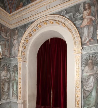 Zdjęcie nr 180: Iluzjonistyczną dekoracją malarską pokryto ściany kaplicy oraz strefę ponadcokołową: ściany tarczowe, pendentywy wokół stiukowych kartuszy herbowych oraz czaszę kopuły i wnętrze latarni. Ściany dolnej części podzielono na dwie strefy. Dolną tworzą zlokalizowane w narożach ścian iluzjonistycznie malowane pary nisz o konchach wypełnionych muszlą. W niszach ukazano osiem stojących frontalnie postaci starców, bez atrybutów i nimbów. Zapewne w ten sposób przedstawiono proroków lub apostołów. Każda z postaci ubrana jest w tunikę oraz płaszcz. Różnicuje je jedynie układ rąk, sposób kształtowania zarostu oraz kolorystyka szat. W drugiej strefie w przyłuczach arkad ukazano całopostaciowe przedstawienia uskrzydlonych aniołów trzymających narzędzia męki Pańskiej. Na ścianie wschodniej anioł prezentuje chustę Weroniki (z prawej) oraz latarnię (z lewej). Po bokach wejścia do kaplicy na ścianie północnej po prawej stronie anioł trzyma lancę i koronę cierniową, a po lewej krzyż. Z kolei na ścianie wschodniej po prawej ukazano anioła z drabiną, a po lewej z gąbką na trzcinie. Natomiast na ścianie południowej, po obu stronach zwieńczenia nagrobka, przedstawiono z prawej anioła z włócznią, a po lewej anioła z wiązką do biczowania. W tej strefie bezpośrednio przy łukach arkad wyobrażono leżące postacie geniuszy malowanych w technice en grisaille. Pod arkadą na ścianie wschodniej (ołtarzowej), ukazano dolną część owalnego jasno ugrowego baldachimu z lambrekinem i zielone, opadające paludamentum, rozsuwane kotarowo przez dwa uskrzydlone anioły. Dekoracja stanowi oprawę dla wiszącego tu obrazu Matki Boskiej z Dzieciątkiem w bogato rzeźbionej ramie. Z kolei na ścianie zachodniej wyobrażono monumentalną scenę Zdjęcia z krzyża. Jest to zwarta kompozycja ogniskująca się na wysokim drewnianym krzyżu, z którego ściągane jest martwe ciało Chrystusa. Z dwóch stron dłuższych ramion krzyż podtrzymują dwaj aniołowie. Ciało Jezusa opuszczane jest na białym pasie tkaniny przewiązanym pod jego ramionami. Chrystus ze zwisającą głową ma ręce ułożone wzdłuż ciała. Biodra przepasane ma białym perizonium. Jego skrzyżowane nogi są ugięte i jeszcze przybite gwoździem do krzyża. Gwóźdź wyciąga z nich obcęgami mężczyzna w niebieskiej, podwiniętej koszuli. Ciało opuszczają dwaj mężczyźni widoczni za poprzecznymi ramionami krzyża, trzymający końce taśmy. Człowiek z lewej strony w niebieskiej koszuli dodatkowo odbiera drugą białą taśmę od mężczyzny ubranego w szaroczarny strój. Osoba ta stoi na drabinie opartej o poprzeczne ramię krzyża. Drugi mężczyzna za krzyżem, w brązowej koszuli, lewą ręką trzyma koniec taśmy, a w uniesionej prawej młotek. Ciało Zbawiciela podtrzymuje lewą ręką kolejny mężczyzna stojący tyłem do widza na drugiej drabinie opartej o lewe ramię krzyża. Jest on ubrany w krótkie brązowe spodenki i bladozieloną koszulę. Pod krzyżem stoi zwarta siedmioosobowa grupa. Najprawdopodobniej z prawej strony ukazano św. Jana i Matkę Boską, na środku dwóch żołnierzy w ostro zakończonych hełmach, dwie kobiety, w tym zapewne Marię Magdalenę i starca widocznego z lewej strony kompozycji. Na ścianach tarczowych nad gzymsem wyobrażono po bokach okrągłych okien siedzących czterech ewangelistów i czterech ojców Kościoła. Na ścianie wschodniej znalazł się z prawej św. Jan z orłem, który w dziobie trzyma złoty kałamarz, a po lewej św. Mateusz rozmawiający z aniołem. Z kolei na ścianie południowej po lewej stronie przedstawiono św. Marka z lwem oraz św. Ambrożego trzymającego w prawej ręce naczynie z wylewającym się miodem. Na ścianie zachodniej z lewej znalazł się czytający św. Augustyn z sercem na dłoni, a z prawej św. Grzegorz Wielki z widoczną za nim tiarą i sową. Na ścianie północnej po lewej ukazano św. Hieronima z leżącym lwem, a po prawej stronie św. Łukasz z wołem. W pendentywach wykonano malarską dekorację w formie wijącej się wstęgi, na tle której widnieją stiukowe kartusze rollwerkowe, o owalnych kaboszonowych polach czwórdzielnych w krzyż. Po stronie wschodniej w pendentywach znajdują się dwa puste kartusze z niezachowanymi herbami. Natomiast herby zachowały się w pendentywach po stronie zachodniej. Na pendentywie północno-zachodnim: Jastrzębiec, Nowina, Gozdawa, Pobóg, a na południowo-zachodnim Gryf, Kotwicz, Kotwicz, Zabawa. W kopule wyobrażono Koronację Matki Boskiej adorowanej przez różnych świętych. Scenę koronacji Marii przez Trójcę Świętą przedstawiono w części wschodniej czaszy. Ukazana frontalnie Maria klęczy ze złożonymi rękami do modlitwy na obłokach. Ma owalną twarz okoloną jasnymi długimi włosami rozsypującymi się na jej ramiona. Na uniesionej ku górze głowie ma ubraną zsuwającą się białą chustę okrywającą włosy. Madonna ubrana jest w zasłaniający jej ramiona bladoniebieski płaszcz, opadający po bokach na obłoki. Nieznacznie wyżej z prawej strony na obłokach siedzi Bóg Ojciec. Wyobrażono go w trzech czwartych w prawo. Jest to starszy mężczyzna o siwych włosach i brodzie. W wyciągniętej prawej ręce nad głową Marii wraz z Chrystusem trzyma złotą koronę w typie zamkniętym. Z kolei w lewej podtrzymuje glob ze złotym krzyżykiem. Bóg Ojciec na głowie ma tiarę papieską. Ubrany jest w szaro-niebieską szatę oraz wzorzystą złotą kapę narzuconą na ramiona i spiętą pod szyją klamrą, z połami okrywającymi nogi. Naprzeciwko siedzi Chrystus widoczny w trzech czwartych w lewo z twarzą ukazaną w profilu. W wyciągniętej prawej ręce trzyma wraz z Bogiem Ojcem koronę, a lewą podtrzymuje złote berło. Chrystus ma jasne włosy opadające puklami na ramiona i podobną krótką brodę. Na głowie ma otwartą złotą koronę. Został przedstawiony z nagim torsem, na którym zaznaczono przebity bok. Jego postać otacza narzucony na ramiona bladoczerwony całun udrapowany w formie płaszcza. Nad koroną bliżej głowy Boga Ojca widoczna jest gołębica Ducha Świętego otoczona świetlistym otokiem. Ponad sceną koronacji wokół otworu do latarni wyobrażono wirujące złocisto-szare obłoki, na tle których widać uskrzydlone główki aniołków oraz postacie mniejszych i większych aniołków, które muzykują na różnych instrumentach, bądź też trzymają gałązki palmowe lub kwiaty albo kwiaty rozsypują. Uskrzydlone główki aniołków widoczne są także pośród obłoków, na których klęczy Madonna. Z prawej strony koronacji wyobrażono dwóch biskupów. Starszy, być może św. Wojciech, został przedstawiony w postawie półklęczącej w trzech czwartych w lewo. Prawą ręką zdaje się wskazywać na scenę koronacji, lewą zaś przyciska do piersi. Ma odkrytą głowę z widocznymi siwymi włosami i taką samą brodą. Ubrany jest w niebiesko-zielonkawą kapę ze złotym kapturem zakończonym chwostem. Na ziemi przy jego stopie widoczna jest złota infuła. Świętemu przyglądają się dwaj aniołowie widoczni z tyłu. Za nim klęczy młodszy biskup z leżącym przy nim na ziemi pastorałem i mieczem. Zapewne jest to św. Stanisław ze Szczepanowa. Święty ukazany został frontalnie z prawą ręką przyciśniętą do piersi, a z lewą nieznacznie odsuniętą w bok. Ma pociągłą twarz o krótkich jasnych włosach i podobnym zaroście. Ubrany jest w albę oraz szaro-bordową kapę o złotym kapturze. Przy jego kolanach stoi złota infuła, a za nim widoczny jest anioł z gałązką palmową. Za św. Stanisławem w rozwianych szatach klęczy św. Jadwiga Śląska, którą identyfikuje bryła kościoła widoczna przy jej stopach. Postać Jadwigi ukazano w profilu z założonymi rękami przed sobą. Na głowie ma niewielką złotą koronę, spod której widoczna jest biała chusta okrywająca włosy. Ubrana jest w bladoczerwoną suknię przepasaną w talii i narzucony na ramiona bladoszkarłatny płaszcz z gronostajowym kołnierzem i obszyciem. Za św. Jadwigą stoi inna święta z odsłoniętą lewą ręką wspartą o bok. Brak atrybutu nie pozwala na jej identyfikację. Ukazano ją w trzech czwartych w prawo z głową w profilu. Ma owalną twarz z wydatnym nosem. Na głowie ma niewielką złotą koronę, spod której widać jasną chustę okrywającą włosy. Ubrana jest w złocisto-szarą suknię z krótkimi rękawami i bladoczerwony rozwiany płaszcz. Pomiędzy obiema kobietami widać dwie postacie uskrzydlonych aniołów. Za świętymi siedzi z otwartą księgą wyobrażony frontalnie św. Jana Kanty. Jest to mężczyzna w średnim wieku z wysokim czołem, krótkimi brązowymi włosami i podobną brodą. Ubrany jest w bladofioletową togę akademicką z gronostajową peleryną. Trzymana przez niego księga ma czerwone okładki. Obok św. Jana Kantego namalowano czteroosobową grupę starszych mężczyzn. Pierwszy z nich, ujętych w trzech czwartych w prawo, zdaje się przyklękając, odbierać prawą ręką palmę męczeństwa podawaną mu przez widocznego nad nim anioła. Lewą ma ułożoną wzdłuż ciała. Ma podłużną twarz widoczną z profilu, otoczoną białymi włosami i spiczastą białą brodą. Ubrany jest w rozwianą jasnobeżową szatę przewiązaną w pasie i taki sam płaszcz narzucony na prawe ramię. Lewy rękaw tuniki jest podciągnięty, odsłaniając przedramię ręki. Na lewej nodze widoczny jest sandał. Za nim przedstawiono klęczącego w ujęciu trzech czwartych w lewo zakonnika eremitę, być może św. Pawła Pustelnika w białym habicie paulińskim. Zakonnik na wysokości bioder ma rozsunięte ręce. Jego twarz o wydatnym nosie otacza krzaczasta, długa, biała broda oraz białe włosy. W głębi za nim częściowo widać dwóch innych brodatych mężczyzn w beżowych szatach. Jeden z nich trzyma w dłoni palmę. W niewielkiej odległości za grupą mężczyzn na pierwszym planie klęczy na obłoku ukazany frontalnie św. Jacek. W rękach uniesionych na wysokości piersi trzyma wieżycową, złotą monstrancję i figurę Matki Boskiej. Dominikanin został przedstawiony z wysokim łysym czołem i wianuszkiem siwiejących włosów. Jego pociągła twarz o wyrazistym nosie i wąskich, nieznacznie zaciśniętych ustach pozbawiona jest zarostu. Ubrany jest w dominikański habit z czarnoniebieskim płaszczem. Z lewej strony sceny koronacji namalowano grupę czterech świętych. Na pierwszym planie widać klęczącego św. Stanisława Kostkę, wspartego na długim kiju z przywiązanym do niego węzełkiem. Święty został przedstawiony w profilu. Ma młodzieńczą, owalną twarz z krótkimi brązowymi włosami i rumianymi policzkami. Ubrany jest w bladobeżowy płaszcz, spod którego widać jasnoseledynową szatę. Za Stanisławem Kostką wyłania się w półpostaci św. Jan Nepomucen z drewnianym krzyżem podtrzymywanym lewą ręką. Jest to dojrzały mężczyzna o pociągłej twarzy, z brązowymi włosami i brodą. Z prawej strony św. Stanisława Kostki ukazano stojące dwie zakonnice, św. Kingę i zapewne bł. Salomeę. Kingę ukazano frontalnie w kontrapoście z głową uniesioną ku niebu w jasnobeżowym habicie klaryski wpadającym w brąz. Księżna prawą ręką przyciska do piersi bryłę soli, a w opuszczonej lewej trzyma kwiat lilii. U jej stóp widać model kościoła klasztornego w Starym Sączu. Obok stoi najprawdopodobniej bł. Salomea. Ukazano ją w mocnym kontrapoście, z dłońmi splecionymi na piersi i spojrzeniem skierowanym ku ziemi. Ubrana jest w rozwiany szaroniebieski habit i jasny welon. Wnętrze latarni wypełniają uskrzydlone głowy aniołków na tle złocisto-szarych obłoków.    







