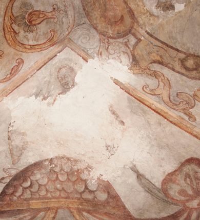 Zdjęcie nr 19: Polichromia pokrywa ściany i sklepienie kolebkowe z lunetami czworobocznej, dwuprzęsłowej zakrystii. Pole sklepienia wypełnia dekoracja ornamentalna utworzona ze zawijanych liści akantu, stylizowanych kwiatów oraz wazonów z kwiatami. Pośrodku sklepienia umieszczono w owalu w kartuszu fragmentarycznie zachowaną scenę Koronacji Marii. W centrum kompozycji zachowała się głowa Marii ujęta w trzy czwarte w prawo z widocznym fragmentem prawego ramienia i ułożonej do modlitwy prawej ręki. Po prawej stronie widać postać tronującego Boga Ojca w złotej koronie, z brodą, trzymającego złotą koronę nad głową Marii. Z lewej można dostrzec fragment ręki Chrystusa oraz fragment jego czerwonej szaty i lewej stopy. Na ścianie wschodniej dekorację malarską w postaci wici utworzonej ze stylizowanych liści akantu i kwiatów poprowadzono w glifie okiennym. Wokół okna utworzono malowane obramienie z motywami stylizowanego ornamentu rollwerkowego. Ponad oknem w glifie znajduje się malowany chrystogram &quot;IHS&quot;. Po prawej stronie okna widoczne jest całopostaciowe przedstawienie proroka Eliasza z rozłożonymi rękami. Postać została ujęta w trzech czwartych w prawo. Ma pociągłą twarz okoloną zarostem z wydatnym nosem i ustami. Ubrana jest w strój wschodni - długą szatę z bladoczerwonym płaszczem z kołnierzykiem spiętym przy szyi oraz z rozciętymi rękawami. Na głowie ma szaroniebieski turban. Ponad nim widać lecącego kruka. Obok mężczyzny z prawej strony widoczna jest niewielka sekwencja krajobrazowa z motywem drzewa. Eliasz zwrócony jest w stronę sceny z Ostatnią Wieczerzą wykonaną w pierwszym przęśle ściany południowej zakrystii. Ostatnią Wieczerzę z prawej strony zamyka malowana kolumna z kapitelem rozdzielająca ścianę na dwa przęsła. Na tle altany ze schodami po lewej, flankowanej trzema kolumnami na wysokich cokołach, ukazano Chrystusa i dwunastu apostołów siedzący przy okrągłym, okazałym stole. Ponad stołem wisi płonącą lampa. Stół nakryto białym obrusem zastawionym siedmioma talerzami, owalną misą pośrodku, trzema okrągłymi bochenkami chleba, dwoma nożami i kielichem. Siedzący centralnie za stołem Chrystus został ukazany frontalnie. Zbawiciel ma zmęczoną twarz otoczoną brodą i długimi włosami, spływającymi na ramiona. Ubrany jest w białą tunikę i niebieski płaszcz przerzucony przez lewe ramię. O jego pierś opiera się zamyślony św. Jan, wspierający głowę na prawej ręce trzymanej na stole. Lewą rękę położoną na blacie ma zgiętą w łokciu. Wokół stołu siedzą rozmawiający ze sobą pozostali apostołowie. Są to starsi, brodaci mężczyźni, niektórzy z nich są łysi. Ubrani są w tuniki i narzucone na ramionach płaszcze. Można spośród nich wyróżnić siedzącego po prawe strony Chrystusa św. Piotra oraz Judasza widocznego jako pierwszego po lewej stronie kompozycji. Judasza ukazanego frontalnie identyfikuje żółta szata oraz trzymany w prawej ręce mieszek ze srebnikami. Powyżej w podłuczu lunety namalowano siedzącego, starszego brodatego mężczyznę ujętego w trzech czwartych w prawo. Jest to ewangelista widoczny do kolan. Prawą rękę przyciska do piersi lewą zaś opiera o stół ustawiony z prawej strony kompozycji z ułożoną na nim księgą. Jego twarz charakteryzuje wydatny nos i usta oraz spojrzenie skierowane na widza. W drugim przęśle ściany  
południowej dekoracje tworzy stylizowana wić akantowo-kwiatowa w glifie okiennym oraz otaczające okno obramienie utworzone z motywów rollwerkowych. Nie zachowała się polichromia w lunecie powyżej okna. Na zachodniej ścianie w glifie dawnego otworu wejściowego obramionego żółtą listwą, wykonano dekorację malarską składającą się z malowanych romboidalnych płycin pokrytych czerwonawą marmoryzacją. Ponad glifem na ścianie znajduje się czerwony krzyż równoramienny rozdzielający datę 16 / 46. Na ścianie z prawej strony otworu wyobrażono Ukrzyżowanie Chrystusa na polu pokrytym licznymi, stojącymi krzyżami. Postać Zbawiciela zachowała się fragmentarycznie w partii tułowia i nóg. Powyżej pole w lunecie ozdobiono stylizowaną wicią akantowo-kwiatową. Ścianę północną w pierwszym przęśle wypełnionym szafą zakrystyjną udekorowano rozpostartym, czerwonym paludamentum z baldachimem. Powyżej w lunecie zachowało się bardzo zniszczone przedstawienie św. Jana Ewangelisty, którego można rozpoznać po młodzieńczej, pociągłej twarz o wydatnych rysach. Z postaci zachowały się jeszcze  fragmenty stóp widoczne nad baldachimem. W drugim prześle w dolnej strefie przepruto drzwi z otworem zamkniętym łukiem odcinkowym, obramienionym złocistą taśmą. W nadprożu znalazła się stylizowana wić akantowo-kwiatowa. Powyżej, w górnej strefie namalowano modlitwę Chrystusa w Ogrójcu. Na pierwszym planie kompozycji widoczny jest klęczący na ziemi Zbawiciel ujęty w trzech czwartych w prawo. Lewą rękę ma przyłożoną do piersi, drugą zaś odsuniętą od tułowia. Jego uniesioną głowę ku górze otacza ciemna broda i długie włosy spływające na plecy. Chrystus ma wydatny nos i usta. Ubrany jest w obszerną, jasnooliwkową tunikę przewiązaną w pasie oraz niebieskoszary płaszcz przerzucony przez lewe ramię i rękę. Jezus wpatruje się w zjawiającego mu się powyżej uskrzydlonego anioła ukazanego w dwóch trzecich na obłokach. Anioł w wyciągniętej prawej ręce trzyma złoty kielich, a prawą podtrzymuje opary o ramię krzyż. Ubrany jest w baldoczerwoną tunikę przewiązaną w pasie. Z prawej strony Jezusa widocznych jest trzech śpiących apostołów. Na pierwszym planie ukazano leżącego św. Piotra z głową wspartą na lewej ręce. Jest to starszy mężczyzna z łysym czołem i siwiejącymi włosami i brodą. Ubrany jest w bladoniebieską tunikę i ciemnoochrowy płaszcz. Za nim widać siedzącego św. Jana z głową wspartą na ręce, którą podpiera o prawe kolano. Jest to młodzieniec o długich włosach, ubrany w blado oliwkową tunikę i baldoczerwony płaszcz. O niewielki wzgórek z lewej strony Jana oparty jest św. Jakub. Jest to starszy mężczyzna z brązową brodą i włosami ubrany w bladooliwkowy płaszcz. Za Zbawicielem z lewej strony kompozycji widać drewniany płot z bramką, przez którą do ogrodu wchodzi Judasz w jasno-chórowej szacie, prowadzący zbrojny oddział sług świątynnych. W głębi kompozycji ukazano panoramę Jerozolimy. Powyżej na sklepieniu lunety wyobrażono siedzącego ewangelistę, który ukazany został jako starzec z długą siwą brodą i łysiejącym czołem. Mężczyzna wspierając prawą rękę o stół podpiera nią głowę. Lewą ma położoną na kolanie. Ubrany jest w szarą tunikę oraz bladoczerwony płaszcz przerzucony przez prawe ramię i okrywający kolana.    
