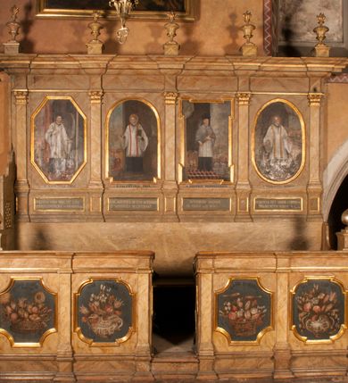 Zdjęcie nr 11: Para przyściennych stalli ustawionych po obu stronach prezbiterium w Niepołomicach uzyskała formę architektoniczną z wysokimi zapleckami i obudowanymi klęcznikami ustawionymi z przodu. Każda z nich otrzymała pięć siedzisk. Wejście do każdej znajduje się z boku i na środku. Przy wejściach z boku na przedpiersiu ustawiono cebulaste sterczyny na prostopadłościennych cokolikach. Zaplecki artykułowano za pomocą pilastrów o kompozytowych kapitelach dźwigających niepełne belkowanie z wydatnym gzymsem, wyłamane na ich osiach. Całość została zwieńczona pięcioma dekoracyjnymi wazami płomienistymi ustawionymi na prostopadłościennych cokolikach. W polach pomiędzy pilastrami umieszczono cztery profilowane, złocone ramy o rożnym kształcie (owalnym, czworobocznym, półkolistym i wielobocznym) z pojedynczymi przedstawieniami  świętych. W stalli południowej od lewej wyobrażono: św. Jana Kantego, Marcina Baryczkę, Świętosława Milczącego, św. Feliksa z Noli. Z kolei w stalli północnej od prawej ukazano: św. Filipa Nereusza, św. Jana Nepomucena, św. Walentego i św. Faustyna z Bresci. Świętych identyfikują czarne, majuskułowe podpisy, których kształt liter sugeruje, że zostały wprowadzone w pierwszej połowie XIX wieku. Poniżej każdego wizerunku na cokołach zaplecków w płycinach wyznaczonych prostokątnymi, złoconymi i profilowanymi ramkami znalazły się łacińskie sentencje. Także lica przedpiersi zostały artykułowane pilastrami na cokołach, jednakże bez dekoracyjnych głowic. Pomiędzy nimi w czworobocznych, profilowanych i złoconych ramach o wklęsłych narożach wyobrażono bukiety kwiatów. Kwiaty ukazano naprzemiennie w płaskich koszach lub wazach na niebieskim tle. Na niektórych bukietach widoczne są siedzące ptaki. Ponad bukietami znalazły się pisane czarnymi literami łacińskie sentencje. Elementy architektoniczne zostały pokryte marmuryzcją, a ramy i detale zdobnicze pozłocono. Na stalli na ścianie północnej ponad kapitelem skrajnego pilastra od zachodu widnieje data ich wykonania: A(NNO) D(OMINI) 1766. Z kolei na stalli na ścianie południowej, ponad kapitelem skrajnego pilastra od zachodu widnieją zapewne inicjały fundatora: X(IĄDZ). / TCD. W / P(LEBANUS) N(IEPOLOMICENSIS).