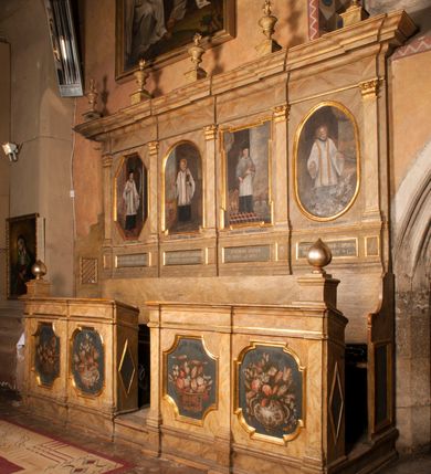 Zdjęcie nr 10: Para przyściennych stalli ustawionych po obu stronach prezbiterium w Niepołomicach uzyskała formę architektoniczną z wysokimi zapleckami i obudowanymi klęcznikami ustawionymi z przodu. Każda z nich otrzymała pięć siedzisk. Wejście do każdej znajduje się z boku i na środku. Przy wejściach z boku na przedpiersiu ustawiono cebulaste sterczyny na prostopadłościennych cokolikach. Zaplecki artykułowano za pomocą pilastrów o kompozytowych kapitelach dźwigających niepełne belkowanie z wydatnym gzymsem, wyłamane na ich osiach. Całość została zwieńczona pięcioma dekoracyjnymi wazami płomienistymi ustawionymi na prostopadłościennych cokolikach. W polach pomiędzy pilastrami umieszczono cztery profilowane, złocone ramy o rożnym kształcie (owalnym, czworobocznym, półkolistym i wielobocznym) z pojedynczymi przedstawieniami  świętych. W stalli południowej od lewej wyobrażono: św. Jana Kantego, Marcina Baryczkę, Świętosława Milczącego, św. Feliksa z Noli. Z kolei w stalli północnej od prawej ukazano: św. Filipa Nereusza, św. Jana Nepomucena, św. Walentego i św. Faustyna z Bresci. Świętych identyfikują czarne, majuskułowe podpisy, których kształt liter sugeruje, że zostały wprowadzone w pierwszej połowie XIX wieku. Poniżej każdego wizerunku na cokołach zaplecków w płycinach wyznaczonych prostokątnymi, złoconymi i profilowanymi ramkami znalazły się łacińskie sentencje. Także lica przedpiersi zostały artykułowane pilastrami na cokołach, jednakże bez dekoracyjnych głowic. Pomiędzy nimi w czworobocznych, profilowanych i złoconych ramach o wklęsłych narożach wyobrażono bukiety kwiatów. Kwiaty ukazano naprzemiennie w płaskich koszach lub wazach na niebieskim tle. Na niektórych bukietach widoczne są siedzące ptaki. Ponad bukietami znalazły się pisane czarnymi literami łacińskie sentencje. Elementy architektoniczne zostały pokryte marmuryzcją, a ramy i detale zdobnicze pozłocono. Na stalli na ścianie północnej ponad kapitelem skrajnego pilastra od zachodu widnieje data ich wykonania: A(NNO) D(OMINI) 1766. Z kolei na stalli na ścianie południowej, ponad kapitelem skrajnego pilastra od zachodu widnieją zapewne inicjały fundatora: X(IĄDZ). / TCD. W / P(LEBANUS) N(IEPOLOMICENSIS).