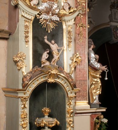 Zdjęcie nr 4: Kamienna chrzcielnica osadzona na wysokim cokole na planie kwadratu, z profilowanym, wielostopniowym trzonem, dekorowanym puklowaniem, liśćmi akantu i uskrzydlonymi główkami anielskimi. Czara puklowana, ozdobiona kartuszem z godłem herbowym Jastrzębiec od frontu oraz kartuszem z godłem herbowym Gryf po przeciwnej stronie, po bokach dwa bukraniony. Chrzcielnica polichromowana w kolorze czerwonym ze złoconymi detalami. Drewniana pokrywa w formie podstawy dekorowanej perełkowaniem i żłobkami, zwieńczona klasycystycznym wazonem zdobionym girlandami, zakończonym kwiatem. 
Chrzcielnica umieszczona w niszy drewnianej oprawy filara arkady tęczowej. Nadbudowa jest architektoniczna, dwukondygnacyjna ze zwieńczeniem. Pierwsza kondygnacja z niszą ujętą dwustrefowym cokołem, zakończonym profilowanym gzymsem dekorowanym żłobkami, zakończonym kartuszem, flankowana kwiatowymi zwisami. Druga kondygnacja z płyciną ujętą pilastrami ozdobionymi kwiatowymi zwisami, zakończonymi gzymsem przechodzącym w woluty, na których znajdują się uskrzydlone główki anielskie. W niszy na rzeźbionym fragmencie skały, flankowanym dwoma drzewami, grupa rzeźbiarska Chrztu Chrystusa. Nad nią na łuku gołębica Ducha Świętego w glorii promienistej. Zwieńczenie w kształcie zbliżonym do kwadratu, ujętego spływami wolutowymi dekorowanymi akantem, zakończone gzymsem i baldachimem. W polu, na obłoku rzeźbiona półpostać Boga Ojca z nimbem trójkątnym i promienistym wokół głowy. Baldachim na planie półkola, w formie niepełnego belkowania, na nim cztery kabłąki podtrzymujące cokół, na którym znajduje się klasycystyczny wazon. Oprawa polichromowana w kolorze seledynowym, ze złoconymi profilami i ornamentyką. 
