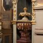 Zdjęcie nr 1: Kamienna chrzcielnica osadzona na wysokim cokole na planie kwadratu, z profilowanym, wielostopniowym trzonem, dekorowanym puklowaniem, liśćmi akantu i uskrzydlonymi główkami anielskimi. Czara puklowana, ozdobiona kartuszem z godłem herbowym Jastrzębiec od frontu oraz kartuszem z godłem herbowym Gryf po przeciwnej stronie, po bokach dwa bukraniony. Chrzcielnica polichromowana w kolorze czerwonym ze złoconymi detalami. Drewniana pokrywa w formie podstawy dekorowanej perełkowaniem i żłobkami, zwieńczona klasycystycznym wazonem zdobionym girlandami, zakończonym kwiatem. 
Chrzcielnica umieszczona w niszy drewnianej oprawy filara arkady tęczowej. Nadbudowa jest architektoniczna, dwukondygnacyjna ze zwieńczeniem. Pierwsza kondygnacja z niszą ujętą dwustrefowym cokołem, zakończonym profilowanym gzymsem dekorowanym żłobkami, zakończonym kartuszem, flankowana kwiatowymi zwisami. Druga kondygnacja z płyciną ujętą pilastrami ozdobionymi kwiatowymi zwisami, zakończonymi gzymsem przechodzącym w woluty, na których znajdują się uskrzydlone główki anielskie. W niszy na rzeźbionym fragmencie skały, flankowanym dwoma drzewami, grupa rzeźbiarska Chrztu Chrystusa. Nad nią na łuku gołębica Ducha Świętego w glorii promienistej. Zwieńczenie w kształcie zbliżonym do kwadratu, ujętego spływami wolutowymi dekorowanymi akantem, zakończone gzymsem i baldachimem. W polu, na obłoku rzeźbiona półpostać Boga Ojca z nimbem trójkątnym i promienistym wokół głowy. Baldachim na planie półkola, w formie niepełnego belkowania, na nim cztery kabłąki podtrzymujące cokół, na którym znajduje się klasycystyczny wazon. Oprawa polichromowana w kolorze seledynowym, ze złoconymi profilami i ornamentyką. 
