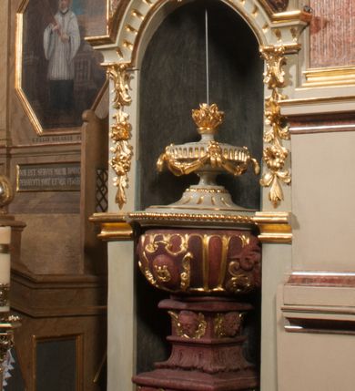 Zdjęcie nr 1: Kamienna chrzcielnica osadzona na wysokim cokole na planie kwadratu, z profilowanym, wielostopniowym trzonem, dekorowanym puklowaniem, liśćmi akantu i uskrzydlonymi główkami anielskimi. Czara puklowana, ozdobiona kartuszem z godłem herbowym Jastrzębiec od frontu oraz kartuszem z godłem herbowym Gryf po przeciwnej stronie, po bokach dwa bukraniony. Chrzcielnica polichromowana w kolorze czerwonym ze złoconymi detalami. Drewniana pokrywa w formie podstawy dekorowanej perełkowaniem i żłobkami, zwieńczona klasycystycznym wazonem zdobionym girlandami, zakończonym kwiatem. 
Chrzcielnica umieszczona w niszy drewnianej oprawy filara arkady tęczowej. Nadbudowa jest architektoniczna, dwukondygnacyjna ze zwieńczeniem. Pierwsza kondygnacja z niszą ujętą dwustrefowym cokołem, zakończonym profilowanym gzymsem dekorowanym żłobkami, zakończonym kartuszem, flankowana kwiatowymi zwisami. Druga kondygnacja z płyciną ujętą pilastrami ozdobionymi kwiatowymi zwisami, zakończonymi gzymsem przechodzącym w woluty, na których znajdują się uskrzydlone główki anielskie. W niszy na rzeźbionym fragmencie skały, flankowanym dwoma drzewami, grupa rzeźbiarska Chrztu Chrystusa. Nad nią na łuku gołębica Ducha Świętego w glorii promienistej. Zwieńczenie w kształcie zbliżonym do kwadratu, ujętego spływami wolutowymi dekorowanymi akantem, zakończone gzymsem i baldachimem. W polu, na obłoku rzeźbiona półpostać Boga Ojca z nimbem trójkątnym i promienistym wokół głowy. Baldachim na planie półkola, w formie niepełnego belkowania, na nim cztery kabłąki podtrzymujące cokół, na którym znajduje się klasycystyczny wazon. Oprawa polichromowana w kolorze seledynowym, ze złoconymi profilami i ornamentyką. 
