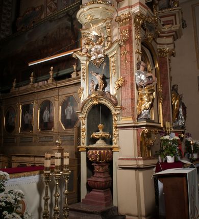Zdjęcie nr 5: Kamienna chrzcielnica osadzona na wysokim cokole na planie kwadratu, z profilowanym, wielostopniowym trzonem, dekorowanym puklowaniem, liśćmi akantu i uskrzydlonymi główkami anielskimi. Czara puklowana, ozdobiona kartuszem z godłem herbowym Jastrzębiec od frontu oraz kartuszem z godłem herbowym Gryf po przeciwnej stronie, po bokach dwa bukraniony. Chrzcielnica polichromowana w kolorze czerwonym ze złoconymi detalami. Drewniana pokrywa w formie podstawy dekorowanej perełkowaniem i żłobkami, zwieńczona klasycystycznym wazonem zdobionym girlandami, zakończonym kwiatem. 
Chrzcielnica umieszczona w niszy drewnianej oprawy filara arkady tęczowej. Nadbudowa jest architektoniczna, dwukondygnacyjna ze zwieńczeniem. Pierwsza kondygnacja z niszą ujętą dwustrefowym cokołem, zakończonym profilowanym gzymsem dekorowanym żłobkami, zakończonym kartuszem, flankowana kwiatowymi zwisami. Druga kondygnacja z płyciną ujętą pilastrami ozdobionymi kwiatowymi zwisami, zakończonymi gzymsem przechodzącym w woluty, na których znajdują się uskrzydlone główki anielskie. W niszy na rzeźbionym fragmencie skały, flankowanym dwoma drzewami, grupa rzeźbiarska Chrztu Chrystusa. Nad nią na łuku gołębica Ducha Świętego w glorii promienistej. Zwieńczenie w kształcie zbliżonym do kwadratu, ujętego spływami wolutowymi dekorowanymi akantem, zakończone gzymsem i baldachimem. W polu, na obłoku rzeźbiona półpostać Boga Ojca z nimbem trójkątnym i promienistym wokół głowy. Baldachim na planie półkola, w formie niepełnego belkowania, na nim cztery kabłąki podtrzymujące cokół, na którym znajduje się klasycystyczny wazon. Oprawa polichromowana w kolorze seledynowym, ze złoconymi profilami i ornamentyką. 
