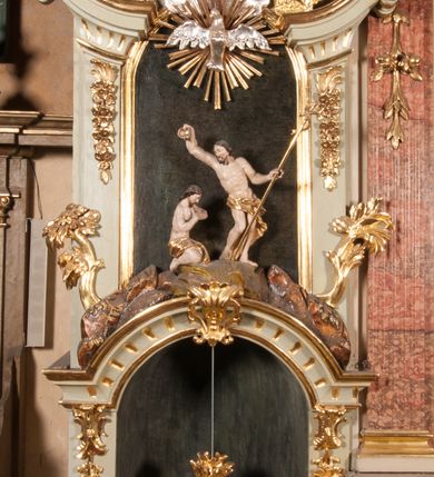 Zdjęcie nr 3: Kamienna chrzcielnica osadzona na wysokim cokole na planie kwadratu, z profilowanym, wielostopniowym trzonem, dekorowanym puklowaniem, liśćmi akantu i uskrzydlonymi główkami anielskimi. Czara puklowana, ozdobiona kartuszem z godłem herbowym Jastrzębiec od frontu oraz kartuszem z godłem herbowym Gryf po przeciwnej stronie, po bokach dwa bukraniony. Chrzcielnica polichromowana w kolorze czerwonym ze złoconymi detalami. Drewniana pokrywa w formie podstawy dekorowanej perełkowaniem i żłobkami, zwieńczona klasycystycznym wazonem zdobionym girlandami, zakończonym kwiatem. 
Chrzcielnica umieszczona w niszy drewnianej oprawy filara arkady tęczowej. Nadbudowa jest architektoniczna, dwukondygnacyjna ze zwieńczeniem. Pierwsza kondygnacja z niszą ujętą dwustrefowym cokołem, zakończonym profilowanym gzymsem dekorowanym żłobkami, zakończonym kartuszem, flankowana kwiatowymi zwisami. Druga kondygnacja z płyciną ujętą pilastrami ozdobionymi kwiatowymi zwisami, zakończonymi gzymsem przechodzącym w woluty, na których znajdują się uskrzydlone główki anielskie. W niszy na rzeźbionym fragmencie skały, flankowanym dwoma drzewami, grupa rzeźbiarska Chrztu Chrystusa. Nad nią na łuku gołębica Ducha Świętego w glorii promienistej. Zwieńczenie w kształcie zbliżonym do kwadratu, ujętego spływami wolutowymi dekorowanymi akantem, zakończone gzymsem i baldachimem. W polu, na obłoku rzeźbiona półpostać Boga Ojca z nimbem trójkątnym i promienistym wokół głowy. Baldachim na planie półkola, w formie niepełnego belkowania, na nim cztery kabłąki podtrzymujące cokół, na którym znajduje się klasycystyczny wazon. Oprawa polichromowana w kolorze seledynowym, ze złoconymi profilami i ornamentyką. 
