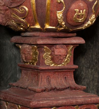 Zdjęcie nr 9: Kamienna chrzcielnica osadzona na wysokim cokole na planie kwadratu, z profilowanym, wielostopniowym trzonem, dekorowanym puklowaniem, liśćmi akantu i uskrzydlonymi główkami anielskimi. Czara puklowana, ozdobiona kartuszem z godłem herbowym Jastrzębiec od frontu oraz kartuszem z godłem herbowym Gryf po przeciwnej stronie, po bokach dwa bukraniony. Chrzcielnica polichromowana w kolorze czerwonym ze złoconymi detalami. Drewniana pokrywa w formie podstawy dekorowanej perełkowaniem i żłobkami, zwieńczona klasycystycznym wazonem zdobionym girlandami, zakończonym kwiatem. 
Chrzcielnica umieszczona w niszy drewnianej oprawy filara arkady tęczowej. Nadbudowa jest architektoniczna, dwukondygnacyjna ze zwieńczeniem. Pierwsza kondygnacja z niszą ujętą dwustrefowym cokołem, zakończonym profilowanym gzymsem dekorowanym żłobkami, zakończonym kartuszem, flankowana kwiatowymi zwisami. Druga kondygnacja z płyciną ujętą pilastrami ozdobionymi kwiatowymi zwisami, zakończonymi gzymsem przechodzącym w woluty, na których znajdują się uskrzydlone główki anielskie. W niszy na rzeźbionym fragmencie skały, flankowanym dwoma drzewami, grupa rzeźbiarska Chrztu Chrystusa. Nad nią na łuku gołębica Ducha Świętego w glorii promienistej. Zwieńczenie w kształcie zbliżonym do kwadratu, ujętego spływami wolutowymi dekorowanymi akantem, zakończone gzymsem i baldachimem. W polu, na obłoku rzeźbiona półpostać Boga Ojca z nimbem trójkątnym i promienistym wokół głowy. Baldachim na planie półkola, w formie niepełnego belkowania, na nim cztery kabłąki podtrzymujące cokół, na którym znajduje się klasycystyczny wazon. Oprawa polichromowana w kolorze seledynowym, ze złoconymi profilami i ornamentyką. 
