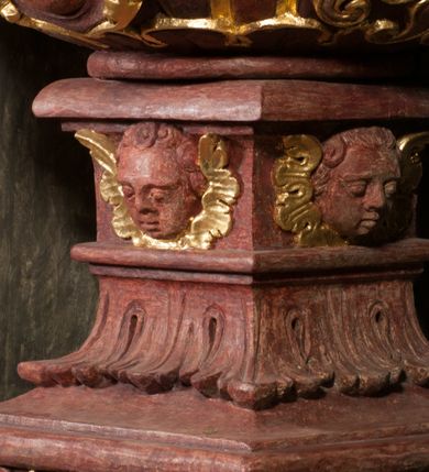 Zdjęcie nr 6: Kamienna chrzcielnica osadzona na wysokim cokole na planie kwadratu, z profilowanym, wielostopniowym trzonem, dekorowanym puklowaniem, liśćmi akantu i uskrzydlonymi główkami anielskimi. Czara puklowana, ozdobiona kartuszem z godłem herbowym Jastrzębiec od frontu oraz kartuszem z godłem herbowym Gryf po przeciwnej stronie, po bokach dwa bukraniony. Chrzcielnica polichromowana w kolorze czerwonym ze złoconymi detalami. Drewniana pokrywa w formie podstawy dekorowanej perełkowaniem i żłobkami, zwieńczona klasycystycznym wazonem zdobionym girlandami, zakończonym kwiatem. 
Chrzcielnica umieszczona w niszy drewnianej oprawy filara arkady tęczowej. Nadbudowa jest architektoniczna, dwukondygnacyjna ze zwieńczeniem. Pierwsza kondygnacja z niszą ujętą dwustrefowym cokołem, zakończonym profilowanym gzymsem dekorowanym żłobkami, zakończonym kartuszem, flankowana kwiatowymi zwisami. Druga kondygnacja z płyciną ujętą pilastrami ozdobionymi kwiatowymi zwisami, zakończonymi gzymsem przechodzącym w woluty, na których znajdują się uskrzydlone główki anielskie. W niszy na rzeźbionym fragmencie skały, flankowanym dwoma drzewami, grupa rzeźbiarska Chrztu Chrystusa. Nad nią na łuku gołębica Ducha Świętego w glorii promienistej. Zwieńczenie w kształcie zbliżonym do kwadratu, ujętego spływami wolutowymi dekorowanymi akantem, zakończone gzymsem i baldachimem. W polu, na obłoku rzeźbiona półpostać Boga Ojca z nimbem trójkątnym i promienistym wokół głowy. Baldachim na planie półkola, w formie niepełnego belkowania, na nim cztery kabłąki podtrzymujące cokół, na którym znajduje się klasycystyczny wazon. Oprawa polichromowana w kolorze seledynowym, ze złoconymi profilami i ornamentyką. 
