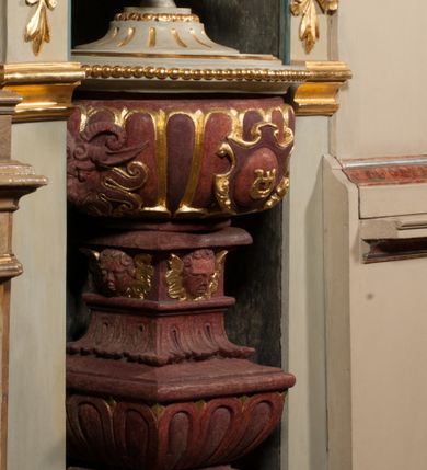 Zdjęcie nr 14: Kamienna chrzcielnica osadzona na wysokim cokole na planie kwadratu, z profilowanym, wielostopniowym trzonem, dekorowanym puklowaniem, liśćmi akantu i uskrzydlonymi główkami anielskimi. Czara puklowana, ozdobiona kartuszem z godłem herbowym Jastrzębiec od frontu oraz kartuszem z godłem herbowym Gryf po przeciwnej stronie, po bokach dwa bukraniony. Chrzcielnica polichromowana w kolorze czerwonym ze złoconymi detalami. Drewniana pokrywa w formie podstawy dekorowanej perełkowaniem i żłobkami, zwieńczona klasycystycznym wazonem zdobionym girlandami, zakończonym kwiatem. 
Chrzcielnica umieszczona w niszy drewnianej oprawy filara arkady tęczowej. Nadbudowa jest architektoniczna, dwukondygnacyjna ze zwieńczeniem. Pierwsza kondygnacja z niszą ujętą dwustrefowym cokołem, zakończonym profilowanym gzymsem dekorowanym żłobkami, zakończonym kartuszem, flankowana kwiatowymi zwisami. Druga kondygnacja z płyciną ujętą pilastrami ozdobionymi kwiatowymi zwisami, zakończonymi gzymsem przechodzącym w woluty, na których znajdują się uskrzydlone główki anielskie. W niszy na rzeźbionym fragmencie skały, flankowanym dwoma drzewami, grupa rzeźbiarska Chrztu Chrystusa. Nad nią na łuku gołębica Ducha Świętego w glorii promienistej. Zwieńczenie w kształcie zbliżonym do kwadratu, ujętego spływami wolutowymi dekorowanymi akantem, zakończone gzymsem i baldachimem. W polu, na obłoku rzeźbiona półpostać Boga Ojca z nimbem trójkątnym i promienistym wokół głowy. Baldachim na planie półkola, w formie niepełnego belkowania, na nim cztery kabłąki podtrzymujące cokół, na którym znajduje się klasycystyczny wazon. Oprawa polichromowana w kolorze seledynowym, ze złoconymi profilami i ornamentyką. 
