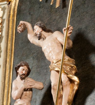 Zdjęcie nr 24: Kamienna chrzcielnica osadzona na wysokim cokole na planie kwadratu, z profilowanym, wielostopniowym trzonem, dekorowanym puklowaniem, liśćmi akantu i uskrzydlonymi główkami anielskimi. Czara puklowana, ozdobiona kartuszem z godłem herbowym Jastrzębiec od frontu oraz kartuszem z godłem herbowym Gryf po przeciwnej stronie, po bokach dwa bukraniony. Chrzcielnica polichromowana w kolorze czerwonym ze złoconymi detalami. Drewniana pokrywa w formie podstawy dekorowanej perełkowaniem i żłobkami, zwieńczona klasycystycznym wazonem zdobionym girlandami, zakończonym kwiatem. 
Chrzcielnica umieszczona w niszy drewnianej oprawy filara arkady tęczowej. Nadbudowa jest architektoniczna, dwukondygnacyjna ze zwieńczeniem. Pierwsza kondygnacja z niszą ujętą dwustrefowym cokołem, zakończonym profilowanym gzymsem dekorowanym żłobkami, zakończonym kartuszem, flankowana kwiatowymi zwisami. Druga kondygnacja z płyciną ujętą pilastrami ozdobionymi kwiatowymi zwisami, zakończonymi gzymsem przechodzącym w woluty, na których znajdują się uskrzydlone główki anielskie. W niszy na rzeźbionym fragmencie skały, flankowanym dwoma drzewami, grupa rzeźbiarska Chrztu Chrystusa. Nad nią na łuku gołębica Ducha Świętego w glorii promienistej. Zwieńczenie w kształcie zbliżonym do kwadratu, ujętego spływami wolutowymi dekorowanymi akantem, zakończone gzymsem i baldachimem. W polu, na obłoku rzeźbiona półpostać Boga Ojca z nimbem trójkątnym i promienistym wokół głowy. Baldachim na planie półkola, w formie niepełnego belkowania, na nim cztery kabłąki podtrzymujące cokół, na którym znajduje się klasycystyczny wazon. Oprawa polichromowana w kolorze seledynowym, ze złoconymi profilami i ornamentyką. 
