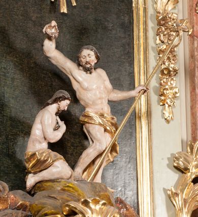 Zdjęcie nr 25: Kamienna chrzcielnica osadzona na wysokim cokole na planie kwadratu, z profilowanym, wielostopniowym trzonem, dekorowanym puklowaniem, liśćmi akantu i uskrzydlonymi główkami anielskimi. Czara puklowana, ozdobiona kartuszem z godłem herbowym Jastrzębiec od frontu oraz kartuszem z godłem herbowym Gryf po przeciwnej stronie, po bokach dwa bukraniony. Chrzcielnica polichromowana w kolorze czerwonym ze złoconymi detalami. Drewniana pokrywa w formie podstawy dekorowanej perełkowaniem i żłobkami, zwieńczona klasycystycznym wazonem zdobionym girlandami, zakończonym kwiatem. 
Chrzcielnica umieszczona w niszy drewnianej oprawy filara arkady tęczowej. Nadbudowa jest architektoniczna, dwukondygnacyjna ze zwieńczeniem. Pierwsza kondygnacja z niszą ujętą dwustrefowym cokołem, zakończonym profilowanym gzymsem dekorowanym żłobkami, zakończonym kartuszem, flankowana kwiatowymi zwisami. Druga kondygnacja z płyciną ujętą pilastrami ozdobionymi kwiatowymi zwisami, zakończonymi gzymsem przechodzącym w woluty, na których znajdują się uskrzydlone główki anielskie. W niszy na rzeźbionym fragmencie skały, flankowanym dwoma drzewami, grupa rzeźbiarska Chrztu Chrystusa. Nad nią na łuku gołębica Ducha Świętego w glorii promienistej. Zwieńczenie w kształcie zbliżonym do kwadratu, ujętego spływami wolutowymi dekorowanymi akantem, zakończone gzymsem i baldachimem. W polu, na obłoku rzeźbiona półpostać Boga Ojca z nimbem trójkątnym i promienistym wokół głowy. Baldachim na planie półkola, w formie niepełnego belkowania, na nim cztery kabłąki podtrzymujące cokół, na którym znajduje się klasycystyczny wazon. Oprawa polichromowana w kolorze seledynowym, ze złoconymi profilami i ornamentyką. 
