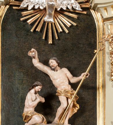 Zdjęcie nr 27: Kamienna chrzcielnica osadzona na wysokim cokole na planie kwadratu, z profilowanym, wielostopniowym trzonem, dekorowanym puklowaniem, liśćmi akantu i uskrzydlonymi główkami anielskimi. Czara puklowana, ozdobiona kartuszem z godłem herbowym Jastrzębiec od frontu oraz kartuszem z godłem herbowym Gryf po przeciwnej stronie, po bokach dwa bukraniony. Chrzcielnica polichromowana w kolorze czerwonym ze złoconymi detalami. Drewniana pokrywa w formie podstawy dekorowanej perełkowaniem i żłobkami, zwieńczona klasycystycznym wazonem zdobionym girlandami, zakończonym kwiatem. 
Chrzcielnica umieszczona w niszy drewnianej oprawy filara arkady tęczowej. Nadbudowa jest architektoniczna, dwukondygnacyjna ze zwieńczeniem. Pierwsza kondygnacja z niszą ujętą dwustrefowym cokołem, zakończonym profilowanym gzymsem dekorowanym żłobkami, zakończonym kartuszem, flankowana kwiatowymi zwisami. Druga kondygnacja z płyciną ujętą pilastrami ozdobionymi kwiatowymi zwisami, zakończonymi gzymsem przechodzącym w woluty, na których znajdują się uskrzydlone główki anielskie. W niszy na rzeźbionym fragmencie skały, flankowanym dwoma drzewami, grupa rzeźbiarska Chrztu Chrystusa. Nad nią na łuku gołębica Ducha Świętego w glorii promienistej. Zwieńczenie w kształcie zbliżonym do kwadratu, ujętego spływami wolutowymi dekorowanymi akantem, zakończone gzymsem i baldachimem. W polu, na obłoku rzeźbiona półpostać Boga Ojca z nimbem trójkątnym i promienistym wokół głowy. Baldachim na planie półkola, w formie niepełnego belkowania, na nim cztery kabłąki podtrzymujące cokół, na którym znajduje się klasycystyczny wazon. Oprawa polichromowana w kolorze seledynowym, ze złoconymi profilami i ornamentyką. 
