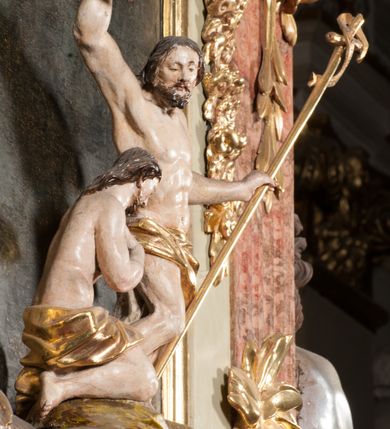 Zdjęcie nr 26: Kamienna chrzcielnica osadzona na wysokim cokole na planie kwadratu, z profilowanym, wielostopniowym trzonem, dekorowanym puklowaniem, liśćmi akantu i uskrzydlonymi główkami anielskimi. Czara puklowana, ozdobiona kartuszem z godłem herbowym Jastrzębiec od frontu oraz kartuszem z godłem herbowym Gryf po przeciwnej stronie, po bokach dwa bukraniony. Chrzcielnica polichromowana w kolorze czerwonym ze złoconymi detalami. Drewniana pokrywa w formie podstawy dekorowanej perełkowaniem i żłobkami, zwieńczona klasycystycznym wazonem zdobionym girlandami, zakończonym kwiatem. 
Chrzcielnica umieszczona w niszy drewnianej oprawy filara arkady tęczowej. Nadbudowa jest architektoniczna, dwukondygnacyjna ze zwieńczeniem. Pierwsza kondygnacja z niszą ujętą dwustrefowym cokołem, zakończonym profilowanym gzymsem dekorowanym żłobkami, zakończonym kartuszem, flankowana kwiatowymi zwisami. Druga kondygnacja z płyciną ujętą pilastrami ozdobionymi kwiatowymi zwisami, zakończonymi gzymsem przechodzącym w woluty, na których znajdują się uskrzydlone główki anielskie. W niszy na rzeźbionym fragmencie skały, flankowanym dwoma drzewami, grupa rzeźbiarska Chrztu Chrystusa. Nad nią na łuku gołębica Ducha Świętego w glorii promienistej. Zwieńczenie w kształcie zbliżonym do kwadratu, ujętego spływami wolutowymi dekorowanymi akantem, zakończone gzymsem i baldachimem. W polu, na obłoku rzeźbiona półpostać Boga Ojca z nimbem trójkątnym i promienistym wokół głowy. Baldachim na planie półkola, w formie niepełnego belkowania, na nim cztery kabłąki podtrzymujące cokół, na którym znajduje się klasycystyczny wazon. Oprawa polichromowana w kolorze seledynowym, ze złoconymi profilami i ornamentyką. 
