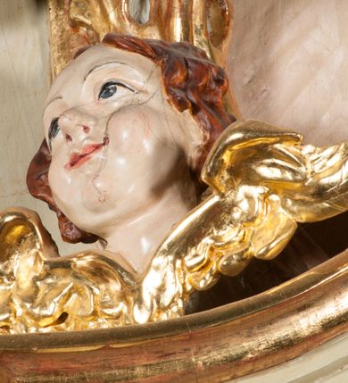 Zdjęcie nr 33: Kamienna chrzcielnica osadzona na wysokim cokole na planie kwadratu, z profilowanym, wielostopniowym trzonem, dekorowanym puklowaniem, liśćmi akantu i uskrzydlonymi główkami anielskimi. Czara puklowana, ozdobiona kartuszem z godłem herbowym Jastrzębiec od frontu oraz kartuszem z godłem herbowym Gryf po przeciwnej stronie, po bokach dwa bukraniony. Chrzcielnica polichromowana w kolorze czerwonym ze złoconymi detalami. Drewniana pokrywa w formie podstawy dekorowanej perełkowaniem i żłobkami, zwieńczona klasycystycznym wazonem zdobionym girlandami, zakończonym kwiatem. 
Chrzcielnica umieszczona w niszy drewnianej oprawy filara arkady tęczowej. Nadbudowa jest architektoniczna, dwukondygnacyjna ze zwieńczeniem. Pierwsza kondygnacja z niszą ujętą dwustrefowym cokołem, zakończonym profilowanym gzymsem dekorowanym żłobkami, zakończonym kartuszem, flankowana kwiatowymi zwisami. Druga kondygnacja z płyciną ujętą pilastrami ozdobionymi kwiatowymi zwisami, zakończonymi gzymsem przechodzącym w woluty, na których znajdują się uskrzydlone główki anielskie. W niszy na rzeźbionym fragmencie skały, flankowanym dwoma drzewami, grupa rzeźbiarska Chrztu Chrystusa. Nad nią na łuku gołębica Ducha Świętego w glorii promienistej. Zwieńczenie w kształcie zbliżonym do kwadratu, ujętego spływami wolutowymi dekorowanymi akantem, zakończone gzymsem i baldachimem. W polu, na obłoku rzeźbiona półpostać Boga Ojca z nimbem trójkątnym i promienistym wokół głowy. Baldachim na planie półkola, w formie niepełnego belkowania, na nim cztery kabłąki podtrzymujące cokół, na którym znajduje się klasycystyczny wazon. Oprawa polichromowana w kolorze seledynowym, ze złoconymi profilami i ornamentyką. 
