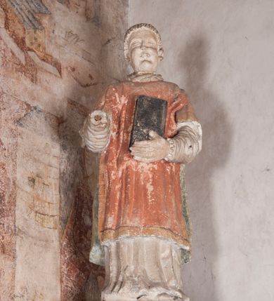 Zdjęcie nr 3: Kamienny lawetarz został osadzony na cokole, którego front zdobi putto trzymające w ręce kartusz herbowy z nieczytelnym godłem. Pod kartuszem widoczna jest czworoboczna wnęka. Nad cokołem znajduje się nisza konchowa z konchą wypełnioną muszlą, ujęta jońskimi pilastrami podtrzymującymi belkowanie, którego fryz ozdobiono główkami aniołków. Archiwolta ponad konchą została na środku przewiązana wolutowym kluczem. Z kolei w przyłuczu konchy znalazły się dwie rozety. Kapitele pilastrów flankujące niszę dodatkowo dekorowane są podwieszonymi girlandami, zaś ich trzony wydłużonymi płycinami. Architraw i gzyms zdobione są kostkowaniem. Na belkowaniu widnieje inskrypcja: „T(HOMAS) O(LINSKI) P(LEBANUS) N(IEPOLOMICENSIS) / A(NNO) D(OMINI) 1696”. Całość wieńczy figura diakona ubranego w białą albę i pomarańczową dalmatykę. W lewej ręce trzyma zamkniętą księgę. Prawa ręka uniesiona na wysokości pasa jest uszkodzona.