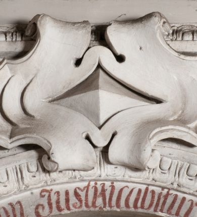 Zdjęcie nr 6: Obraz uzyskał kształt stojącego prostokąta zamkniętego łukiem pełnym. Na tle komnaty o brązowych ścianach, z widoczną po prawej stronie arkadą otwierającą się na kolejne pomieszczenie, ukazany został św. Karol Boromeusz rozdający jałmużnę ubogim. Świętego przedstawiono w całej postaci, zwróconego w trzech czwartych w prawo. Lewą ręką sięga po pieniądze z zielonej szkatuły leżącej na stole obok, prawą natomiast wyciąga w kierunku ubogich. Ze szkatuły wysuwa się purpurowa chusta. Święty Karol Boromeusz ma owalną twarz o prostym nosie, lekko uśmiechniętych ustach, półprzymkniętych oczach, okoloną ciemnobrązowymi, krótkimi włosami. Ubrany jest w strój kardynalski: purpurową sutannę, mucet i piuskę oraz białą komżę. Jego głowę otacza świetlisty nimb. Po jego lewej stronie widoczny jest stół, przykryty sięgającym ziemi białym obrusem ze złotymi frędzlami. Ustawiono na nim drewniany krucyfiks, księgę oraz wspomnianą zieloną szkatułę. Za postacią świętego widoczny jest ozdobny fotel o wysokim zaplecku z zielonym obiciem. Po prawej stronie świętego w wejściu do komnaty przedstawiono w całej postaci żebraków. Pierwszy mężczyzna zwrócony jest profilem w lewo. Wyciąga prawą rękę po pieniądze podawane mu przez świętego, wspierając się lewą dłonią na kosturze. Ma twarz o ciemnobrązowej karnacji, okoloną ciemną, długą brodą. Głowę ma przewiązaną białą chustą. Postać ubrana jest w brunatną tunikę sięgającą kolan oraz czarne buty. Drugi mężczyzna, wspierający się na kosturze, zwrócony jest w trzech czwartych w lewo. Niewidoczna jest jego lewa ręka. Lewą nogę ma uciętą poniżej kolana i zabandażowaną. Powyżej kolana ma owiniętą białym opatrunkiem prawą nogę. Jego twarz jest owalna o ciemnobrązowej karnacji. Przedstawiony został boso w grynszpanowej tunice przewiązanej w pasie i sięgającej kolan, odsłaniającej prawe ramię. Powyżej sceny namalowano podwieszoną, złoto-umbrową kotarę ze złotymi frędzlami.
Ponad obrazem na archiwolcie arkady znajduje się czerwony łaciński napis: „Qui aurum diligit, non iustisificabitur Eccles[iastes] Cap[ut] 31 vers[us] 5”. Natomiast na dolnej krawędzi wnęki napis polski, także wykonany rubrą: „Złoto ubogim Karol ze szkatuły daye / znać ze złotem do nieba nikt się nie dostaye”.