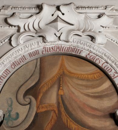 Zdjęcie nr 5: Obraz uzyskał kształt stojącego prostokąta zamkniętego łukiem pełnym. Na tle komnaty o brązowych ścianach, z widoczną po prawej stronie arkadą otwierającą się na kolejne pomieszczenie, ukazany został św. Karol Boromeusz rozdający jałmużnę ubogim. Świętego przedstawiono w całej postaci, zwróconego w trzech czwartych w prawo. Lewą ręką sięga po pieniądze z zielonej szkatuły leżącej na stole obok, prawą natomiast wyciąga w kierunku ubogich. Ze szkatuły wysuwa się purpurowa chusta. Święty Karol Boromeusz ma owalną twarz o prostym nosie, lekko uśmiechniętych ustach, półprzymkniętych oczach, okoloną ciemnobrązowymi, krótkimi włosami. Ubrany jest w strój kardynalski: purpurową sutannę, mucet i piuskę oraz białą komżę. Jego głowę otacza świetlisty nimb. Po jego lewej stronie widoczny jest stół, przykryty sięgającym ziemi białym obrusem ze złotymi frędzlami. Ustawiono na nim drewniany krucyfiks, księgę oraz wspomnianą zieloną szkatułę. Za postacią świętego widoczny jest ozdobny fotel o wysokim zaplecku z zielonym obiciem. Po prawej stronie świętego w wejściu do komnaty przedstawiono w całej postaci żebraków. Pierwszy mężczyzna zwrócony jest profilem w lewo. Wyciąga prawą rękę po pieniądze podawane mu przez świętego, wspierając się lewą dłonią na kosturze. Ma twarz o ciemnobrązowej karnacji, okoloną ciemną, długą brodą. Głowę ma przewiązaną białą chustą. Postać ubrana jest w brunatną tunikę sięgającą kolan oraz czarne buty. Drugi mężczyzna, wspierający się na kosturze, zwrócony jest w trzech czwartych w lewo. Niewidoczna jest jego lewa ręka. Lewą nogę ma uciętą poniżej kolana i zabandażowaną. Powyżej kolana ma owiniętą białym opatrunkiem prawą nogę. Jego twarz jest owalna o ciemnobrązowej karnacji. Przedstawiony został boso w grynszpanowej tunice przewiązanej w pasie i sięgającej kolan, odsłaniającej prawe ramię. Powyżej sceny namalowano podwieszoną, złoto-umbrową kotarę ze złotymi frędzlami.
Ponad obrazem na archiwolcie arkady znajduje się czerwony łaciński napis: „Qui aurum diligit, non iustisificabitur Eccles[iastes] Cap[ut] 31 vers[us] 5”. Natomiast na dolnej krawędzi wnęki napis polski, także wykonany rubrą: „Złoto ubogim Karol ze szkatuły daye / znać ze złotem do nieba nikt się nie dostaye”.