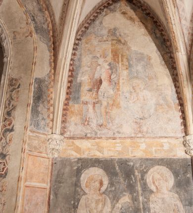 Zdjęcie nr 13: Zachowana dekoracja malarska znajduje się w tzw. starej zakrystii, określanej także jako oratorium królewskie. Dobudowano ją od północy do prezbiterium niepołomickiego kościoła. Jest to niewielka budowla na rzucie wydłużonego prostokąta, nakryta trójprzęsłowym krzyżowo-żebrowym sklepieniem, od wschodu zamknięta dwubocznie. Od zachodu w narożniku południowym widoczne jest załamanie muru, tworzącego dwie ściany wieżyczki schodowej. Do jej wnętrza prowadzi ostrołukowy portal. Oratorium oświetlają trzy ostrołukowe okna. Tylko czwarte, obecnie zamurowane, przeprute w murze północno-wschodnim pochodzi z czasów budowy kościoła. Stara zakrystia połączona jest z kościołem za pomocą ostrołukowego, profilowanego kamiennego portalu umieszczonego w środkowym przęśle ściany południowej. Na sklepieniu znajdują się rzeźbione zworniki z przedstawieniem w przęśle wschodnim głowy Chrystusa w nimbie krzyżowym, w środkowym maski liściastej, a w zachodnim motywów roślinnych. Podobnie rzeźbione motywy roślinne  pokrywają wsporniki, na które spływają żebra. 
Zachowana polichromia tworzy z architekturą starej zakrystii harmonijną całość. Białe wysklepki sklepień otoczone są wielobarwnymi, namalowanymi wzdłuż żeber, iluzjonistyczno-geometrycznymi pasami imitującymi kamienne inkrustacje w typie &quot;arte cosmatesca&quot;. Pierwotnie polichromowane były także same żebra oraz rzeźbione zworniki i wsporniki. Pionowym rytmom barwnych pasów przeciwstawiają się na ścianie wschodniej, południowej i zachodniej dwa poziome, iluzjonistyczne malowane fryzy. Górny, w odcieniach brązów jest szerszy i przypomina gzyms. Przebiega w połowie wysokości ścian, na poziomie wsporników. Poniżej znajduje się drugi, o połowę węższy, skomponowany z wielobarwnych geometrycznych rozet w rombowych polach. Fryzy wyznaczają trzy strefy dekoracji. Dolny pas uległ zniszczeniu za wyjątkiem niewielkiego fragmentu iluzjonistycznie malowanej konsolki na ścianie południowej. Dla strefy środkowej głównej tło stanowi niebieska barwa. Niestety oba te pasy obecnie są mocno uszkodzone. 
Pierwotnie pas środkowy ścian zajmowały naturalnej wielkości postacie kobiece, ujęte powyżej kolan. Z namalowanych dwunastu świętych do naszych czasów przetrwało sześć - w całości cztery (na ścianie wschodniej, południowej i zachodniej) oraz jedna bez głowy (ściana południowa) i jedna w połowie (ściana zachodnia). Są to jasno lub ciemnowłose młode kobiety, trzymające palmy męczeństwa. Ubrane są w suknie z narzuconymi płaszczami w kolorach żółtym, oliwkowym, różowym, liliowym, brązowym i błękitnym. Jedna z nich, wyobrażona ponad wejściem na klatkę schodową w wieżyczce, jest ukazana jako kobieta w podeszłym wieku z okrytą głową i dłońmi uniesionymi na wysokość piersi w geście orantki. Głowy męczennic i orantki okalają plastycznie, tłoczone w zaprawie nimby zdobione promieniami. Na niektórych głowach dziewic zachowały się ślady namalowanych brązową farbą koron o niskich sterczynach w kształcie heraldycznych lilii. 
W ścianie północno-wschodniej dwubocznego zamknięcia znajduje się obecnie zamurowane lancetowate, ostrołukowe okno z fragmentami polichromii w glifie okiennym. Pierwotnie tworzyło ją dziesięć czterolistnych obramowań wpisanych w romby, umieszczonych jeden nad drugim. Przedstawiono w nich w półpostaci aniołów z głowami otoczonymi nimbami. W dolnej części okno do  poziomu gotyckich wsporników otacza iluzjonistycznie malowana dekoracja utworzona z motywów kamiennych płyt.  
W górnej strefie oratorium wyobrażono cykl hagiograficzny, składający się z sześciu scen namalowanych w ostrołukowych polach, wyznaczonych łukami tarczowymi sklepień i ujętych w obramienia z plastycznie malowanych zębów. Ograniczający je od dołu, iluzjonistycznie malowany, jakby wystający gzyms nadaje przedstawieniom teatralną głębie. 
Na ścianie południowo-wschodniej przedstawiono bliżej niezidentyfikowaną scenę męczeństwa. Na tle ochrowo-żółtej architektury ukazano z lewej strony na pierwszym planie grupę  rycerzy, z których dwóch widocznych jest w całej postaci w średniowiecznych zbrojach stylizowanych na rzymskie, w hełmach i z ostrołukowymi tarczami trzymanymi w lewych rękach. Ich odzież ma kolor błękitny, granatowy, czerwony, fioletowy, cynobrowy. Rycerze lekko unoszą głowy w stronę półpostaci wychylającej się z okna loggi w górnej części architektury. Był to zapewne władca, dziś jego postać zachowała się tylko fragmentarycznie. Postać z okna wskazuje palcem na centralnie ukazany kocioł zawieszony pod arkadą, stanowiącą część architektury tła. Widoczna w nim jest w półpostaci jasnowłosa kobieta w białej sukni na ramiączkach z modlitewnie złożonymi rękami na piersi. Jej głowę otacza nimb. Kocioł wisi na haku za półokrągły uchwyt, pod nim pali się ognisko. Z prawej strony kotła widoczna jest fragmentarycznie zachowana postać rycerza w zbroi, który oburącz długim kijem miesza w kotle. 
Na prawo od sceny męczeństwa na ścianie południowej namalowano scenę określaną jako &quot;przed władcą&quot;. W centrum przedstawienia na tle architektury wyobrażono tron z zasiadającym na nim władcą. Jest to brodaty mężczyzna ukazany z profilu z koroną na głowie, który w prawej dłoni unosi berło skierowane w stronę przyprowadzonej do niego kobiety. Tron i architektoniczne jego obramienie malowane jest w kolorach brązu i ochry. Jego zaplecek dekoruje zawieszona pasiasta tkanina. Ciemnowłosy władca ubrany jest w turkusową szatę z narzuconym na ramiona brązowym płaszczem. Za tronem z lewej strony stoi paź o kędzierzawej fryzurze wsparty na trzymanym ostrzem w dół mieczu. Stojąca przed władcą kobieta ubrana jest w czerwoną długą suknię. Ma jasne włosy, a jej głowę okala nimb. Ręce ma skrzyżowane i związane na wysokości pasa. Obok niej stoi inna postać także w długiej szacie, z uniesioną prawą dłonią. Za świętą widoczne są inne postacie, niestety skala zniszczenia malowidła uniemożliwia ich opis i identyfikację. 
Trzecie malowidło, usytuowane nad wejściem do oratorium, ukazuje scenę dysputyt. W centrum przedstawienia pod architektonicznym zadaszeniem w formie portyku lub altany o kasetonowym stropie, wspartym na dwóch czworobocznych filarach, ukazano widoczne z profilu trzy siedzące  kobiety. Jedna z lewej strony a dwie naprzeciwko z prawej. Kobieta z lewej  ma odkryte jasne, długie włosy. Jej głowę otacza nimb. Na wysokość piersi unosi obie dłonie, sprawiając wrażenie, jakby w nich trzymała owalny przedmiot. Ubrana jest w ciemnoczerwoną długą suknię, zakrywającą nogi. Naprzeciw niej siedzą dwie inne niewiasty o długich, jasnych włosach w prostych koronach na głowach. Jedna z nich unosi swoją prawą rękę. Obie ubrane są w długie suknie w kolorze ochry. Za nimi pomiędzy filarami widoczna jest rozpięta zasłona w pionowe oliwkowe, zielone, białe i czerwone pasy. Na podłodze leży dywan dekorowany w żółte, zielone, niebieskie i czerwone pasy. 
Mocno zniszczone w dolnej części czwarte przedstawienie ilustruje scenę chłosty. Z lewej strony kompozycji wyobrażono budowlę z architektoniczną loggią, z której w półpostaci wychyla się władca. W prawej ręce poziomo trzyma berło skierowane w stronę grupy osób, za którymi wykonywana jest kara chłosty. Władca, podobnie jak w drugiej scenie, został ukazany jako mężczyzna o ciemnym zaroście i takich samych włosach. Na głowie ma koronę. Ubrany jest w brązowe szaty o różnych odcieniach. Centralnie przedstawioną stłoczoną grupę tworzy czterech żołnierzy oraz starszy mężczyzna w zawoju na głowie z siwą, długą brodą. Żołnierze mają zbroje i hełmy. Jeden z nich, najbliżej stojący z lewej przedstawiony został w profilu z głową i prawą ręką skierowaną w stronę króla. Także w stronę władcy zwrócony jest mężczyzna w zawoju na głowie z dwiema dłońmi uniesionymi na wysokość piersi. Z prawej strony kompozycji namalowano, dziś fragmentarycznie zachowany, rodzaj drewnianej szubienicy, utworzonej z poprzecznie zawieszonego na drewnianych podporach drążka. Nad dróżką do góry nogami wisi postać (dziś zniszczona), z której zachowały się jedynie stopy związane w kostkach sznurem. W jej stronę zwraca się jeden z żołnierzy ze środkowej grupy. Po prawej stronie szubienicy widoczna jest część postaci oprawcy, z której zachował się tylko fragment głowy oraz uniesiona lewa ręka z drewnianą pałką. 
Dwie ostatnie sceny umieszczono na zachodnim murze oratorium. Pierwsza z nich przedstawia strącenie bożka i została połączona z drugą ukazującą uwolnienia z więzienia. Część centralną kompozycji stanowi potrzaskana, wywracająca się kolumna, z której spada szatan w postaci żywego demona pokrytego sierścią. Strącił go widoczny w półpostaci w górnej części malowidła anioł trzymający w prawej ręce miecz. Z lewej strony kompozycji  klęczy na skalistej ziemi święta ubrana w długą ciemnoczerwoną suknię. Niestety nie zachowała się górna część jej postaci. Za nią stoi stłoczona grupa przerażonych świadków tego wydarzenia. Natomiast z prawej strony malowidła wyobrażono scenę uwolnienia z więzienia. Jej zasadniczym elementem jest architektoniczna struktura nawiązująca kształtem do wieży mieszkalnej, więzienia bądź bramy miejskiej. Święta ze związanymi rękami w ciemnoczerwonej sukni wychodzi z niej z prawej strony. W centrum budowli widoczne jest półokrągłe okno, a w nim zapewne jej popiersie o długich jasnych włosach, trzymające w dłoniach jakiś przedmiot. Wszystkie sceny cyklu w jedną całość wiąże gładkie niebieskie tło. Niezachowana kompozycja polichromii na ścianie północnej musiała odbiegać swym układem od pozostałych ścian. Płaszczyzna ścian została podzielona za pomocą węższego dolnego fryzu na dwie strefy. Górną część najprawdopodobniej zajmowały duże postacie stojące (fragment jednej z nich zachował się w sąsiedztwie okna w ścianie północno-wschodniej). Pod fryzem w części dolnej najprawdopodobniej była namalowana iluzjonistycznie wisząca kotara.   
    




