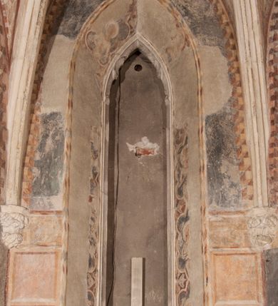 Zdjęcie nr 15: Zachowana dekoracja malarska znajduje się w tzw. starej zakrystii, określanej także jako oratorium królewskie. Dobudowano ją od północy do prezbiterium niepołomickiego kościoła. Jest to niewielka budowla na rzucie wydłużonego prostokąta, nakryta trójprzęsłowym krzyżowo-żebrowym sklepieniem, od wschodu zamknięta dwubocznie. Od zachodu w narożniku południowym widoczne jest załamanie muru, tworzącego dwie ściany wieżyczki schodowej. Do jej wnętrza prowadzi ostrołukowy portal. Oratorium oświetlają trzy ostrołukowe okna. Tylko czwarte, obecnie zamurowane, przeprute w murze północno-wschodnim pochodzi z czasów budowy kościoła. Stara zakrystia połączona jest z kościołem za pomocą ostrołukowego, profilowanego kamiennego portalu umieszczonego w środkowym przęśle ściany południowej. Na sklepieniu znajdują się rzeźbione zworniki z przedstawieniem w przęśle wschodnim głowy Chrystusa w nimbie krzyżowym, w środkowym maski liściastej, a w zachodnim motywów roślinnych. Podobnie rzeźbione motywy roślinne  pokrywają wsporniki, na które spływają żebra. 
Zachowana polichromia tworzy z architekturą starej zakrystii harmonijną całość. Białe wysklepki sklepień otoczone są wielobarwnymi, namalowanymi wzdłuż żeber, iluzjonistyczno-geometrycznymi pasami imitującymi kamienne inkrustacje w typie &quot;arte cosmatesca&quot;. Pierwotnie polichromowane były także same żebra oraz rzeźbione zworniki i wsporniki. Pionowym rytmom barwnych pasów przeciwstawiają się na ścianie wschodniej, południowej i zachodniej dwa poziome, iluzjonistyczne malowane fryzy. Górny, w odcieniach brązów jest szerszy i przypomina gzyms. Przebiega w połowie wysokości ścian, na poziomie wsporników. Poniżej znajduje się drugi, o połowę węższy, skomponowany z wielobarwnych geometrycznych rozet w rombowych polach. Fryzy wyznaczają trzy strefy dekoracji. Dolny pas uległ zniszczeniu za wyjątkiem niewielkiego fragmentu iluzjonistycznie malowanej konsolki na ścianie południowej. Dla strefy środkowej głównej tło stanowi niebieska barwa. Niestety oba te pasy obecnie są mocno uszkodzone. 
Pierwotnie pas środkowy ścian zajmowały naturalnej wielkości postacie kobiece, ujęte powyżej kolan. Z namalowanych dwunastu świętych do naszych czasów przetrwało sześć - w całości cztery (na ścianie wschodniej, południowej i zachodniej) oraz jedna bez głowy (ściana południowa) i jedna w połowie (ściana zachodnia). Są to jasno lub ciemnowłose młode kobiety, trzymające palmy męczeństwa. Ubrane są w suknie z narzuconymi płaszczami w kolorach żółtym, oliwkowym, różowym, liliowym, brązowym i błękitnym. Jedna z nich, wyobrażona ponad wejściem na klatkę schodową w wieżyczce, jest ukazana jako kobieta w podeszłym wieku z okrytą głową i dłońmi uniesionymi na wysokość piersi w geście orantki. Głowy męczennic i orantki okalają plastycznie, tłoczone w zaprawie nimby zdobione promieniami. Na niektórych głowach dziewic zachowały się ślady namalowanych brązową farbą koron o niskich sterczynach w kształcie heraldycznych lilii. 
W ścianie północno-wschodniej dwubocznego zamknięcia znajduje się obecnie zamurowane lancetowate, ostrołukowe okno z fragmentami polichromii w glifie okiennym. Pierwotnie tworzyło ją dziesięć czterolistnych obramowań wpisanych w romby, umieszczonych jeden nad drugim. Przedstawiono w nich w półpostaci aniołów z głowami otoczonymi nimbami. W dolnej części okno do  poziomu gotyckich wsporników otacza iluzjonistycznie malowana dekoracja utworzona z motywów kamiennych płyt.  
W górnej strefie oratorium wyobrażono cykl hagiograficzny, składający się z sześciu scen namalowanych w ostrołukowych polach, wyznaczonych łukami tarczowymi sklepień i ujętych w obramienia z plastycznie malowanych zębów. Ograniczający je od dołu, iluzjonistycznie malowany, jakby wystający gzyms nadaje przedstawieniom teatralną głębie. 
Na ścianie południowo-wschodniej przedstawiono bliżej niezidentyfikowaną scenę męczeństwa. Na tle ochrowo-żółtej architektury ukazano z lewej strony na pierwszym planie grupę  rycerzy, z których dwóch widocznych jest w całej postaci w średniowiecznych zbrojach stylizowanych na rzymskie, w hełmach i z ostrołukowymi tarczami trzymanymi w lewych rękach. Ich odzież ma kolor błękitny, granatowy, czerwony, fioletowy, cynobrowy. Rycerze lekko unoszą głowy w stronę półpostaci wychylającej się z okna loggi w górnej części architektury. Był to zapewne władca, dziś jego postać zachowała się tylko fragmentarycznie. Postać z okna wskazuje palcem na centralnie ukazany kocioł zawieszony pod arkadą, stanowiącą część architektury tła. Widoczna w nim jest w półpostaci jasnowłosa kobieta w białej sukni na ramiączkach z modlitewnie złożonymi rękami na piersi. Jej głowę otacza nimb. Kocioł wisi na haku za półokrągły uchwyt, pod nim pali się ognisko. Z prawej strony kotła widoczna jest fragmentarycznie zachowana postać rycerza w zbroi, który oburącz długim kijem miesza w kotle. 
Na prawo od sceny męczeństwa na ścianie południowej namalowano scenę określaną jako &quot;przed władcą&quot;. W centrum przedstawienia na tle architektury wyobrażono tron z zasiadającym na nim władcą. Jest to brodaty mężczyzna ukazany z profilu z koroną na głowie, który w prawej dłoni unosi berło skierowane w stronę przyprowadzonej do niego kobiety. Tron i architektoniczne jego obramienie malowane jest w kolorach brązu i ochry. Jego zaplecek dekoruje zawieszona pasiasta tkanina. Ciemnowłosy władca ubrany jest w turkusową szatę z narzuconym na ramiona brązowym płaszczem. Za tronem z lewej strony stoi paź o kędzierzawej fryzurze wsparty na trzymanym ostrzem w dół mieczu. Stojąca przed władcą kobieta ubrana jest w czerwoną długą suknię. Ma jasne włosy, a jej głowę okala nimb. Ręce ma skrzyżowane i związane na wysokości pasa. Obok niej stoi inna postać także w długiej szacie, z uniesioną prawą dłonią. Za świętą widoczne są inne postacie, niestety skala zniszczenia malowidła uniemożliwia ich opis i identyfikację. 
Trzecie malowidło, usytuowane nad wejściem do oratorium, ukazuje scenę dysputyt. W centrum przedstawienia pod architektonicznym zadaszeniem w formie portyku lub altany o kasetonowym stropie, wspartym na dwóch czworobocznych filarach, ukazano widoczne z profilu trzy siedzące  kobiety. Jedna z lewej strony a dwie naprzeciwko z prawej. Kobieta z lewej  ma odkryte jasne, długie włosy. Jej głowę otacza nimb. Na wysokość piersi unosi obie dłonie, sprawiając wrażenie, jakby w nich trzymała owalny przedmiot. Ubrana jest w ciemnoczerwoną długą suknię, zakrywającą nogi. Naprzeciw niej siedzą dwie inne niewiasty o długich, jasnych włosach w prostych koronach na głowach. Jedna z nich unosi swoją prawą rękę. Obie ubrane są w długie suknie w kolorze ochry. Za nimi pomiędzy filarami widoczna jest rozpięta zasłona w pionowe oliwkowe, zielone, białe i czerwone pasy. Na podłodze leży dywan dekorowany w żółte, zielone, niebieskie i czerwone pasy. 
Mocno zniszczone w dolnej części czwarte przedstawienie ilustruje scenę chłosty. Z lewej strony kompozycji wyobrażono budowlę z architektoniczną loggią, z której w półpostaci wychyla się władca. W prawej ręce poziomo trzyma berło skierowane w stronę grupy osób, za którymi wykonywana jest kara chłosty. Władca, podobnie jak w drugiej scenie, został ukazany jako mężczyzna o ciemnym zaroście i takich samych włosach. Na głowie ma koronę. Ubrany jest w brązowe szaty o różnych odcieniach. Centralnie przedstawioną stłoczoną grupę tworzy czterech żołnierzy oraz starszy mężczyzna w zawoju na głowie z siwą, długą brodą. Żołnierze mają zbroje i hełmy. Jeden z nich, najbliżej stojący z lewej przedstawiony został w profilu z głową i prawą ręką skierowaną w stronę króla. Także w stronę władcy zwrócony jest mężczyzna w zawoju na głowie z dwiema dłońmi uniesionymi na wysokość piersi. Z prawej strony kompozycji namalowano, dziś fragmentarycznie zachowany, rodzaj drewnianej szubienicy, utworzonej z poprzecznie zawieszonego na drewnianych podporach drążka. Nad dróżką do góry nogami wisi postać (dziś zniszczona), z której zachowały się jedynie stopy związane w kostkach sznurem. W jej stronę zwraca się jeden z żołnierzy ze środkowej grupy. Po prawej stronie szubienicy widoczna jest część postaci oprawcy, z której zachował się tylko fragment głowy oraz uniesiona lewa ręka z drewnianą pałką. 
Dwie ostatnie sceny umieszczono na zachodnim murze oratorium. Pierwsza z nich przedstawia strącenie bożka i została połączona z drugą ukazującą uwolnienia z więzienia. Część centralną kompozycji stanowi potrzaskana, wywracająca się kolumna, z której spada szatan w postaci żywego demona pokrytego sierścią. Strącił go widoczny w półpostaci w górnej części malowidła anioł trzymający w prawej ręce miecz. Z lewej strony kompozycji  klęczy na skalistej ziemi święta ubrana w długą ciemnoczerwoną suknię. Niestety nie zachowała się górna część jej postaci. Za nią stoi stłoczona grupa przerażonych świadków tego wydarzenia. Natomiast z prawej strony malowidła wyobrażono scenę uwolnienia z więzienia. Jej zasadniczym elementem jest architektoniczna struktura nawiązująca kształtem do wieży mieszkalnej, więzienia bądź bramy miejskiej. Święta ze związanymi rękami w ciemnoczerwonej sukni wychodzi z niej z prawej strony. W centrum budowli widoczne jest półokrągłe okno, a w nim zapewne jej popiersie o długich jasnych włosach, trzymające w dłoniach jakiś przedmiot. Wszystkie sceny cyklu w jedną całość wiąże gładkie niebieskie tło. Niezachowana kompozycja polichromii na ścianie północnej musiała odbiegać swym układem od pozostałych ścian. Płaszczyzna ścian została podzielona za pomocą węższego dolnego fryzu na dwie strefy. Górną część najprawdopodobniej zajmowały duże postacie stojące (fragment jednej z nich zachował się w sąsiedztwie okna w ścianie północno-wschodniej). Pod fryzem w części dolnej najprawdopodobniej była namalowana iluzjonistycznie wisząca kotara.   
    




