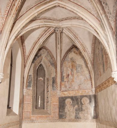 Zdjęcie nr 16: Zachowana dekoracja malarska znajduje się w tzw. starej zakrystii, określanej także jako oratorium królewskie. Dobudowano ją od północy do prezbiterium niepołomickiego kościoła. Jest to niewielka budowla na rzucie wydłużonego prostokąta, nakryta trójprzęsłowym krzyżowo-żebrowym sklepieniem, od wschodu zamknięta dwubocznie. Od zachodu w narożniku południowym widoczne jest załamanie muru, tworzącego dwie ściany wieżyczki schodowej. Do jej wnętrza prowadzi ostrołukowy portal. Oratorium oświetlają trzy ostrołukowe okna. Tylko czwarte, obecnie zamurowane, przeprute w murze północno-wschodnim pochodzi z czasów budowy kościoła. Stara zakrystia połączona jest z kościołem za pomocą ostrołukowego, profilowanego kamiennego portalu umieszczonego w środkowym przęśle ściany południowej. Na sklepieniu znajdują się rzeźbione zworniki z przedstawieniem w przęśle wschodnim głowy Chrystusa w nimbie krzyżowym, w środkowym maski liściastej, a w zachodnim motywów roślinnych. Podobnie rzeźbione motywy roślinne  pokrywają wsporniki, na które spływają żebra. 
Zachowana polichromia tworzy z architekturą starej zakrystii harmonijną całość. Białe wysklepki sklepień otoczone są wielobarwnymi, namalowanymi wzdłuż żeber, iluzjonistyczno-geometrycznymi pasami imitującymi kamienne inkrustacje w typie &quot;arte cosmatesca&quot;. Pierwotnie polichromowane były także same żebra oraz rzeźbione zworniki i wsporniki. Pionowym rytmom barwnych pasów przeciwstawiają się na ścianie wschodniej, południowej i zachodniej dwa poziome, iluzjonistyczne malowane fryzy. Górny, w odcieniach brązów jest szerszy i przypomina gzyms. Przebiega w połowie wysokości ścian, na poziomie wsporników. Poniżej znajduje się drugi, o połowę węższy, skomponowany z wielobarwnych geometrycznych rozet w rombowych polach. Fryzy wyznaczają trzy strefy dekoracji. Dolny pas uległ zniszczeniu za wyjątkiem niewielkiego fragmentu iluzjonistycznie malowanej konsolki na ścianie południowej. Dla strefy środkowej głównej tło stanowi niebieska barwa. Niestety oba te pasy obecnie są mocno uszkodzone. 
Pierwotnie pas środkowy ścian zajmowały naturalnej wielkości postacie kobiece, ujęte powyżej kolan. Z namalowanych dwunastu świętych do naszych czasów przetrwało sześć - w całości cztery (na ścianie wschodniej, południowej i zachodniej) oraz jedna bez głowy (ściana południowa) i jedna w połowie (ściana zachodnia). Są to jasno lub ciemnowłose młode kobiety, trzymające palmy męczeństwa. Ubrane są w suknie z narzuconymi płaszczami w kolorach żółtym, oliwkowym, różowym, liliowym, brązowym i błękitnym. Jedna z nich, wyobrażona ponad wejściem na klatkę schodową w wieżyczce, jest ukazana jako kobieta w podeszłym wieku z okrytą głową i dłońmi uniesionymi na wysokość piersi w geście orantki. Głowy męczennic i orantki okalają plastycznie, tłoczone w zaprawie nimby zdobione promieniami. Na niektórych głowach dziewic zachowały się ślady namalowanych brązową farbą koron o niskich sterczynach w kształcie heraldycznych lilii. 
W ścianie północno-wschodniej dwubocznego zamknięcia znajduje się obecnie zamurowane lancetowate, ostrołukowe okno z fragmentami polichromii w glifie okiennym. Pierwotnie tworzyło ją dziesięć czterolistnych obramowań wpisanych w romby, umieszczonych jeden nad drugim. Przedstawiono w nich w półpostaci aniołów z głowami otoczonymi nimbami. W dolnej części okno do  poziomu gotyckich wsporników otacza iluzjonistycznie malowana dekoracja utworzona z motywów kamiennych płyt.  
W górnej strefie oratorium wyobrażono cykl hagiograficzny, składający się z sześciu scen namalowanych w ostrołukowych polach, wyznaczonych łukami tarczowymi sklepień i ujętych w obramienia z plastycznie malowanych zębów. Ograniczający je od dołu, iluzjonistycznie malowany, jakby wystający gzyms nadaje przedstawieniom teatralną głębie. 
Na ścianie południowo-wschodniej przedstawiono bliżej niezidentyfikowaną scenę męczeństwa. Na tle ochrowo-żółtej architektury ukazano z lewej strony na pierwszym planie grupę  rycerzy, z których dwóch widocznych jest w całej postaci w średniowiecznych zbrojach stylizowanych na rzymskie, w hełmach i z ostrołukowymi tarczami trzymanymi w lewych rękach. Ich odzież ma kolor błękitny, granatowy, czerwony, fioletowy, cynobrowy. Rycerze lekko unoszą głowy w stronę półpostaci wychylającej się z okna loggi w górnej części architektury. Był to zapewne władca, dziś jego postać zachowała się tylko fragmentarycznie. Postać z okna wskazuje palcem na centralnie ukazany kocioł zawieszony pod arkadą, stanowiącą część architektury tła. Widoczna w nim jest w półpostaci jasnowłosa kobieta w białej sukni na ramiączkach z modlitewnie złożonymi rękami na piersi. Jej głowę otacza nimb. Kocioł wisi na haku za półokrągły uchwyt, pod nim pali się ognisko. Z prawej strony kotła widoczna jest fragmentarycznie zachowana postać rycerza w zbroi, który oburącz długim kijem miesza w kotle. 
Na prawo od sceny męczeństwa na ścianie południowej namalowano scenę określaną jako &quot;przed władcą&quot;. W centrum przedstawienia na tle architektury wyobrażono tron z zasiadającym na nim władcą. Jest to brodaty mężczyzna ukazany z profilu z koroną na głowie, który w prawej dłoni unosi berło skierowane w stronę przyprowadzonej do niego kobiety. Tron i architektoniczne jego obramienie malowane jest w kolorach brązu i ochry. Jego zaplecek dekoruje zawieszona pasiasta tkanina. Ciemnowłosy władca ubrany jest w turkusową szatę z narzuconym na ramiona brązowym płaszczem. Za tronem z lewej strony stoi paź o kędzierzawej fryzurze wsparty na trzymanym ostrzem w dół mieczu. Stojąca przed władcą kobieta ubrana jest w czerwoną długą suknię. Ma jasne włosy, a jej głowę okala nimb. Ręce ma skrzyżowane i związane na wysokości pasa. Obok niej stoi inna postać także w długiej szacie, z uniesioną prawą dłonią. Za świętą widoczne są inne postacie, niestety skala zniszczenia malowidła uniemożliwia ich opis i identyfikację. 
Trzecie malowidło, usytuowane nad wejściem do oratorium, ukazuje scenę dysputyt. W centrum przedstawienia pod architektonicznym zadaszeniem w formie portyku lub altany o kasetonowym stropie, wspartym na dwóch czworobocznych filarach, ukazano widoczne z profilu trzy siedzące  kobiety. Jedna z lewej strony a dwie naprzeciwko z prawej. Kobieta z lewej  ma odkryte jasne, długie włosy. Jej głowę otacza nimb. Na wysokość piersi unosi obie dłonie, sprawiając wrażenie, jakby w nich trzymała owalny przedmiot. Ubrana jest w ciemnoczerwoną długą suknię, zakrywającą nogi. Naprzeciw niej siedzą dwie inne niewiasty o długich, jasnych włosach w prostych koronach na głowach. Jedna z nich unosi swoją prawą rękę. Obie ubrane są w długie suknie w kolorze ochry. Za nimi pomiędzy filarami widoczna jest rozpięta zasłona w pionowe oliwkowe, zielone, białe i czerwone pasy. Na podłodze leży dywan dekorowany w żółte, zielone, niebieskie i czerwone pasy. 
Mocno zniszczone w dolnej części czwarte przedstawienie ilustruje scenę chłosty. Z lewej strony kompozycji wyobrażono budowlę z architektoniczną loggią, z której w półpostaci wychyla się władca. W prawej ręce poziomo trzyma berło skierowane w stronę grupy osób, za którymi wykonywana jest kara chłosty. Władca, podobnie jak w drugiej scenie, został ukazany jako mężczyzna o ciemnym zaroście i takich samych włosach. Na głowie ma koronę. Ubrany jest w brązowe szaty o różnych odcieniach. Centralnie przedstawioną stłoczoną grupę tworzy czterech żołnierzy oraz starszy mężczyzna w zawoju na głowie z siwą, długą brodą. Żołnierze mają zbroje i hełmy. Jeden z nich, najbliżej stojący z lewej przedstawiony został w profilu z głową i prawą ręką skierowaną w stronę króla. Także w stronę władcy zwrócony jest mężczyzna w zawoju na głowie z dwiema dłońmi uniesionymi na wysokość piersi. Z prawej strony kompozycji namalowano, dziś fragmentarycznie zachowany, rodzaj drewnianej szubienicy, utworzonej z poprzecznie zawieszonego na drewnianych podporach drążka. Nad dróżką do góry nogami wisi postać (dziś zniszczona), z której zachowały się jedynie stopy związane w kostkach sznurem. W jej stronę zwraca się jeden z żołnierzy ze środkowej grupy. Po prawej stronie szubienicy widoczna jest część postaci oprawcy, z której zachował się tylko fragment głowy oraz uniesiona lewa ręka z drewnianą pałką. 
Dwie ostatnie sceny umieszczono na zachodnim murze oratorium. Pierwsza z nich przedstawia strącenie bożka i została połączona z drugą ukazującą uwolnienia z więzienia. Część centralną kompozycji stanowi potrzaskana, wywracająca się kolumna, z której spada szatan w postaci żywego demona pokrytego sierścią. Strącił go widoczny w półpostaci w górnej części malowidła anioł trzymający w prawej ręce miecz. Z lewej strony kompozycji  klęczy na skalistej ziemi święta ubrana w długą ciemnoczerwoną suknię. Niestety nie zachowała się górna część jej postaci. Za nią stoi stłoczona grupa przerażonych świadków tego wydarzenia. Natomiast z prawej strony malowidła wyobrażono scenę uwolnienia z więzienia. Jej zasadniczym elementem jest architektoniczna struktura nawiązująca kształtem do wieży mieszkalnej, więzienia bądź bramy miejskiej. Święta ze związanymi rękami w ciemnoczerwonej sukni wychodzi z niej z prawej strony. W centrum budowli widoczne jest półokrągłe okno, a w nim zapewne jej popiersie o długich jasnych włosach, trzymające w dłoniach jakiś przedmiot. Wszystkie sceny cyklu w jedną całość wiąże gładkie niebieskie tło. Niezachowana kompozycja polichromii na ścianie północnej musiała odbiegać swym układem od pozostałych ścian. Płaszczyzna ścian została podzielona za pomocą węższego dolnego fryzu na dwie strefy. Górną część najprawdopodobniej zajmowały duże postacie stojące (fragment jednej z nich zachował się w sąsiedztwie okna w ścianie północno-wschodniej). Pod fryzem w części dolnej najprawdopodobniej była namalowana iluzjonistycznie wisząca kotara.   
    




